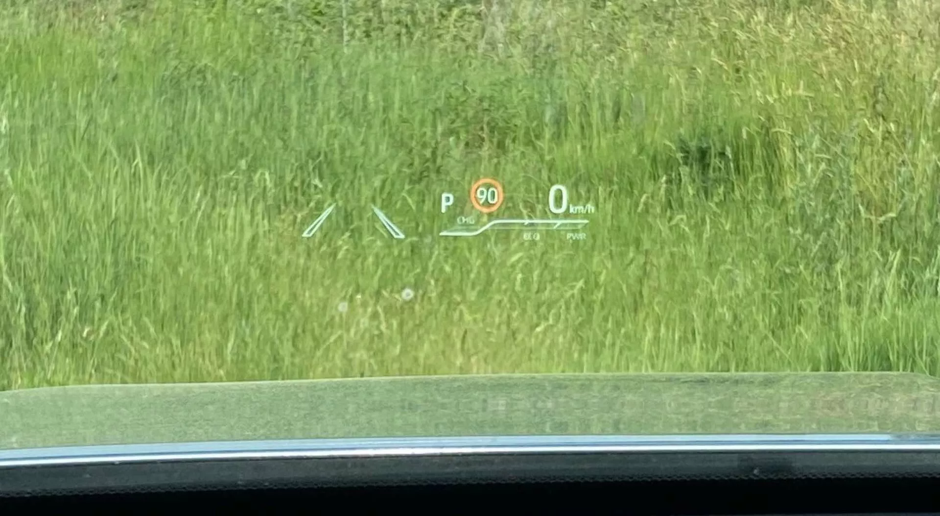 La vue intérieure d'une Toyota Highlander 2021 2.5 Hybrid Business Edition se concentre sur l'affichage tête haute (HUD) projeté sur le pare-brise avant. Le HUD affiche des informations de conduite essentielles : un indicateur de vitesse 'P' pour le 'Park', un panneau de limitation de vitesse '90', une vitesse actuelle de '0 km/h' indiquant que la voiture est à l'arrêt et des symboles possiblement liés à un système d'assistance au stationnement ou de maintien de voie. La scène est prise légèrement à droite du siège du conducteur, donnant sur une zone herbeuse à l'extérieur.