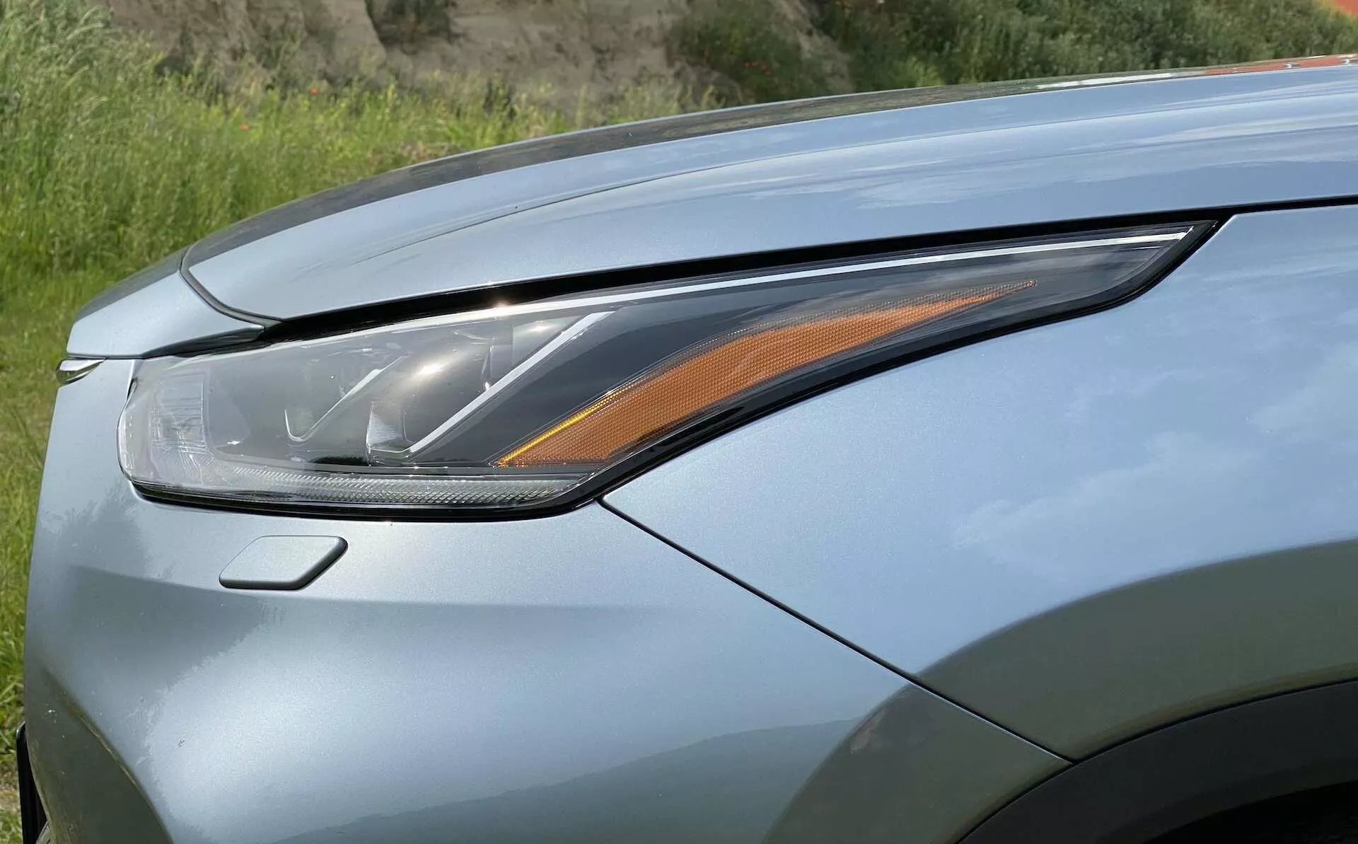 Gros plan de la section avant gauche d'un Toyota Highlander 2.5 Hybrid Business Edition 2021, mettant en avant le bloc optique et une partie du capot. L'image est prise d'un angle bas, légèrement vers le haut et vers l'avant, soulignant le phare moderne à LED ou laser avec plusieurs éléments lumineux, y compris le clignotant ambre. Le capot présente une peinture métallique avec une finition nacrée possible et des lignes aérodynamiques, mettant en avant la technologie d’éclairage avancée et le design luxueux.