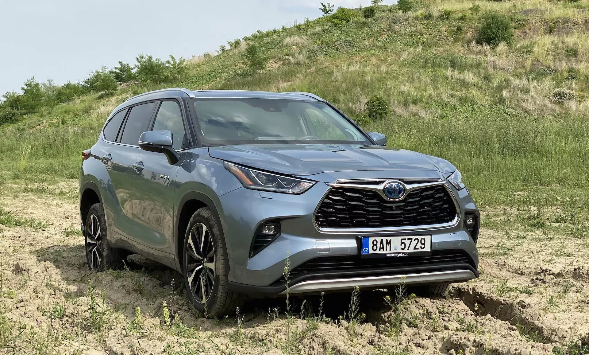 Vue de trois quarts avant d'un SUV Toyota Highlander 2.5 Hybrid Business Edition gris, positionné en extérieur dans une zone hors route avec de l'herbe et de la terre. L'image met en évidence la calandre avant proéminente avec l'emblème de la marque, les phares LED distinctifs qui s'étendent sur les côtés, les jantes en alliage multi-branches d'environ 18-20 pouces, les lignes sculptées de la carrosserie avec des ailes galbées, la hauteur surélevée suggérant des capacités de traction intégrale, les feux de brouillard du pare-chocs avant, les rétroviseurs latéraux avec indicateurs de clignotant intégrés et les vitres teintées à l'arrière. L'image offre une vue claire de ces caractéristiques à hauteur d'œil, avec une lumière naturelle montrant les détails de couleur et de design de la voiture.