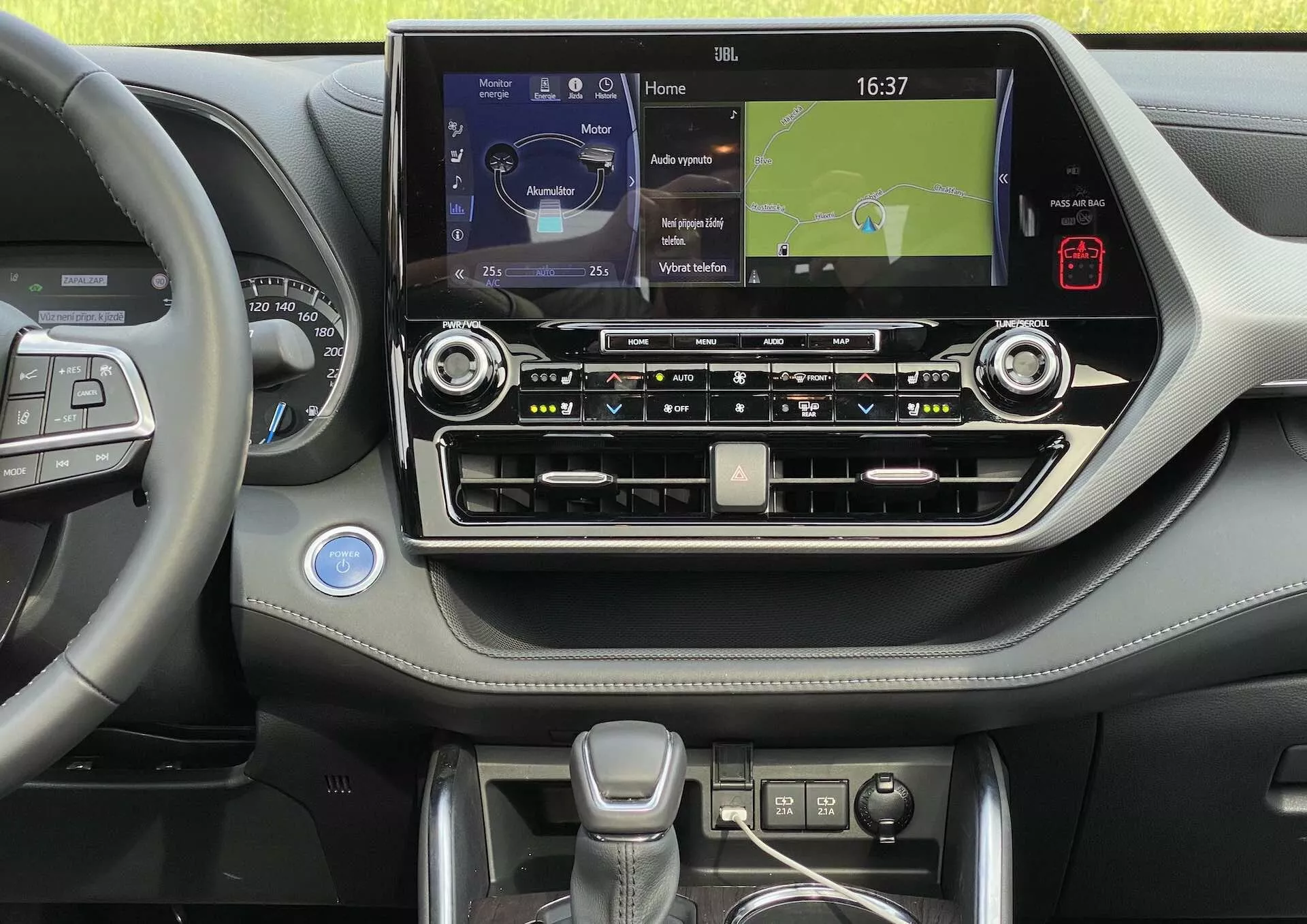 L'intérieur d'une Toyota Highlander 2021, se concentrant sur la console centrale et une partie du côté conducteur. L'image, prise à hauteur des yeux, révèle le bord du volant avec des boutons multifonctions, suggérant des fonctionnalités telles que le régulateur de vitesse, le contrôle du système audio et la gestion des appels mains libres. La console centrale dispose d'un grand écran tactile affichant une carte de navigation, indiquant la connectivité des médias et du téléphone. Sous l'écran tactile, il y a des boutons et des boutons pour les systèmes de climatisation et audio, ainsi qu'une horloge analogique, des accents métalliques et un tableau de bord en cuir ou doux au toucher. Le levier de vitesses est moderne et ergonomique, avec des commandes supplémentaires autour de lui, probablement pour les modes de conduite. En bas, on aperçoit des porte-gobelets ou un espace de rangement. L'intérieur est propre, bien éclairé et bien entretenu.