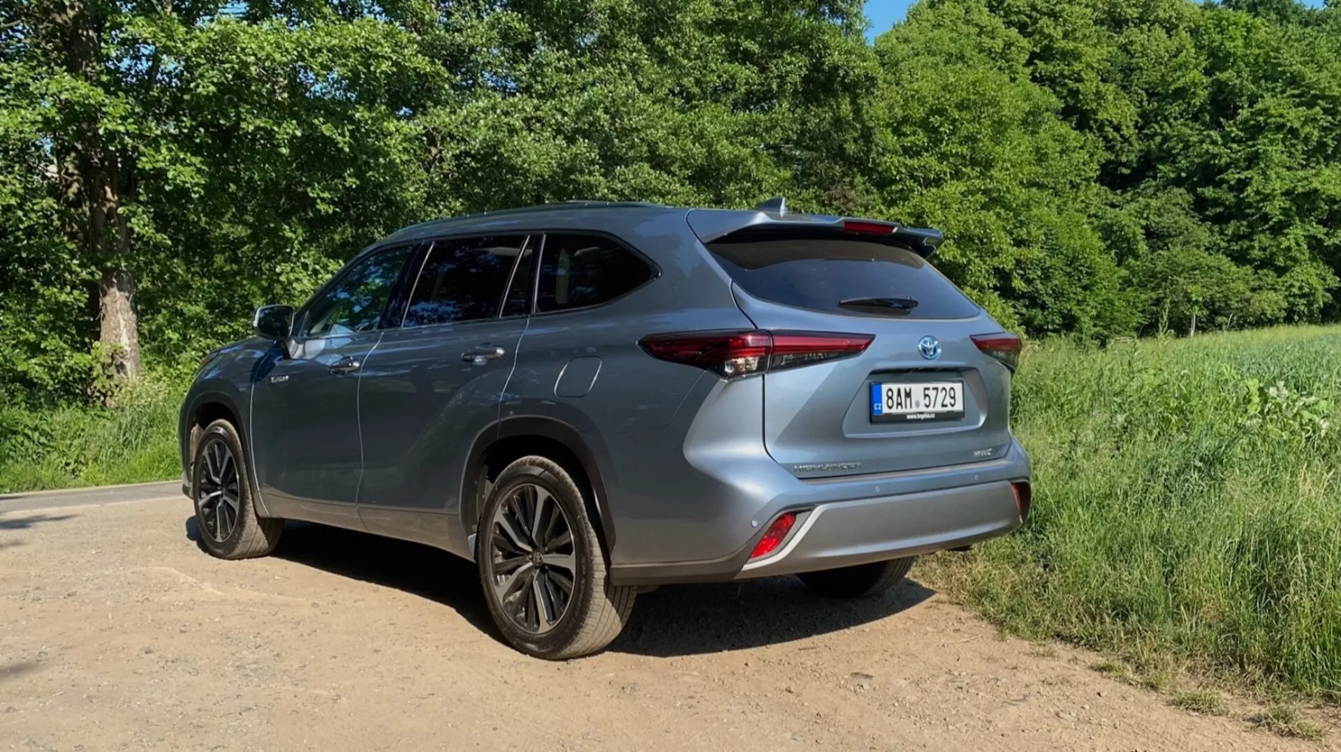 Toyota Highlander 2021 Hybrid Business Edition présenté sous un angle arrière trois-quarts, mettant en valeur le côté droit et l'arrière de la voiture. La prise de vue au niveau du sol met en évidence les roues en alliage foncé et la hauteur globale. Le design arrière, avec des feux arrière élégants, un pare-chocs sculpté et un hayon avec un spoiler arrière, est accentué. L'image montre également le design des portes, des fenêtres latérales et des rétroviseurs. Le véhicule est positionné sur un chemin de terre ou de gravier avec un feuillage vert et des arbres en arrière-plan. L'éclairage ensoleillé projette des ombres vers l'avant, suggérant que le soleil est derrière la caméra.