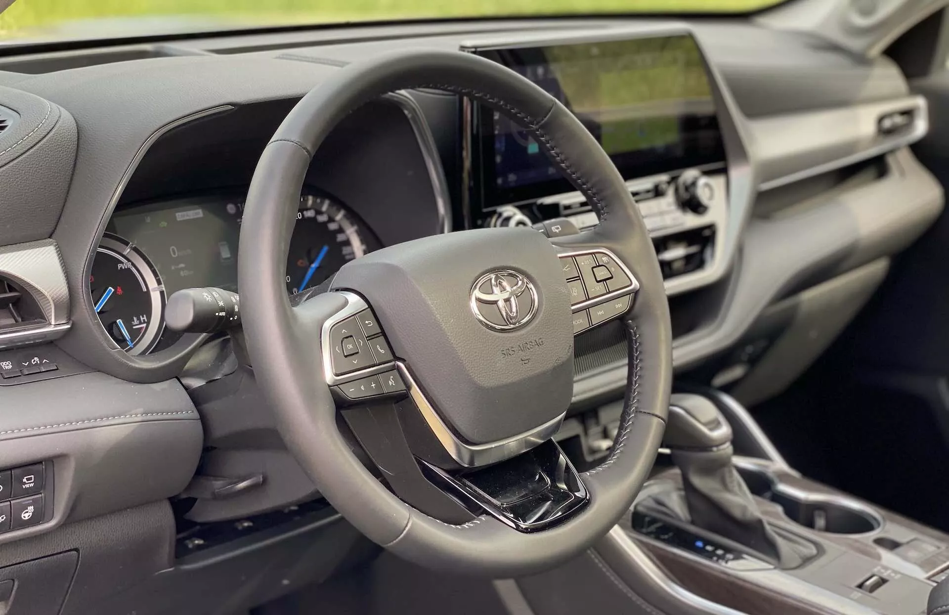 Vue intérieure d'une Toyota Highlander 2021 2.5 Hybrid Business Edition, mettant l'accent sur le côté conducteur. L'image, prise d'un angle frontal droit, présente en évidence l'emblème Toyota, les boutons multimédia et d'affichage des informations à gauche, et les boutons du régulateur de vitesse adaptatif et de communication mains libres à droite. Le tableau de bord derrière le volant inclut un affichage numérique à thème bleu et deux jauges analogiques, indiquant un véhicule hybride. Le tableau de bord central comprend une unité d'infodivertissement tactile intégrée, des bouches d'aération et des commandes de climatisation. Les matériaux de l'habitacle suggèrent un design moderne avec fonctionnalité et luxe, avec des garnitures en cuir ou similicuir, un levier de changement de vitesse métallique pour la transmission automatique et divers contrôles de conduite. La lumière naturelle illumine l'intérieur, mettant en valeur les détails et les textures autour du volant et du tableau de bord.