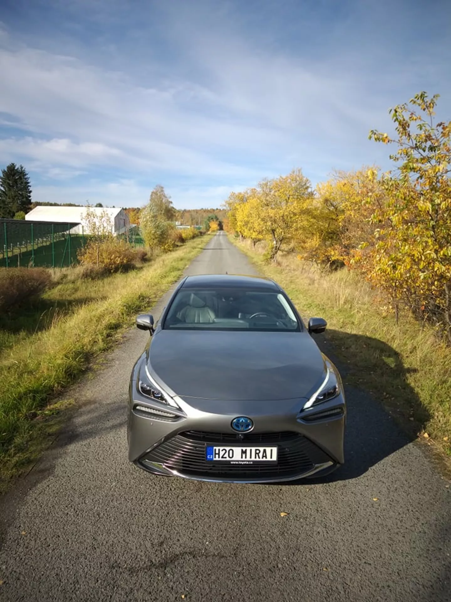 Toyota Mirai 2021 photographiée de face sous un angle légèrement élevé, mettant en vedette ses phares LED élancés et sa grille proéminente avec un écusson central pouvant inclure la technologie des capteurs, dans un cadre semi-rural sous la lumière naturelle, démontrant la technologie des cellules à combustible à hydrogène avancée et la conception aérodynamique