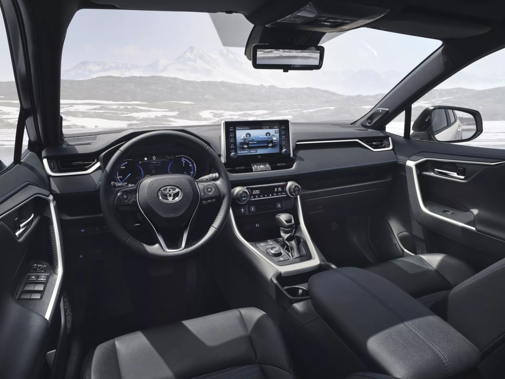 Vue intérieure du Toyota RAV4 2019 depuis le siège arrière, mettant en avant le design moderne et les fonctionnalités technologiques incluant le volant avec boutons de contrôle, affichage numérique, système d'infodivertissement tactile, levier de vitesse automatique, sièges tapissés en foncé, et commandes sur le panneau de porte avec un paysage montagneux enneigé visible à travers le pare-brise.