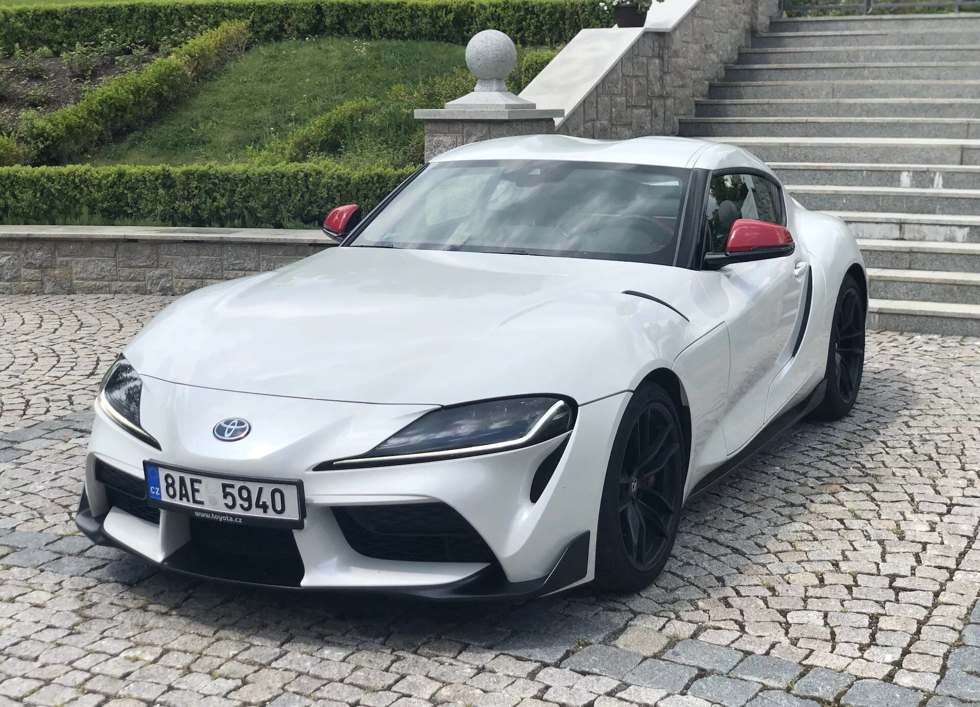 L'image montre une Toyota Supra 2019 (toyota-gr-supra-2-0-pure-automatic) blanche, prise sous un angle trois quarts de face. La posture basse et large de la voiture, les phares angulaires et agressifs, ainsi que les jantes en alliage multi-branches noires sont bien visibles. La photo met en valeur le design élégant, les contours aérodynamiques et l'esthétique de performance. La voiture de sport est garée sur le trottoir avec un bord et un escalier en arrière-plan, suggérant un environnement urbain ou résidentiel.