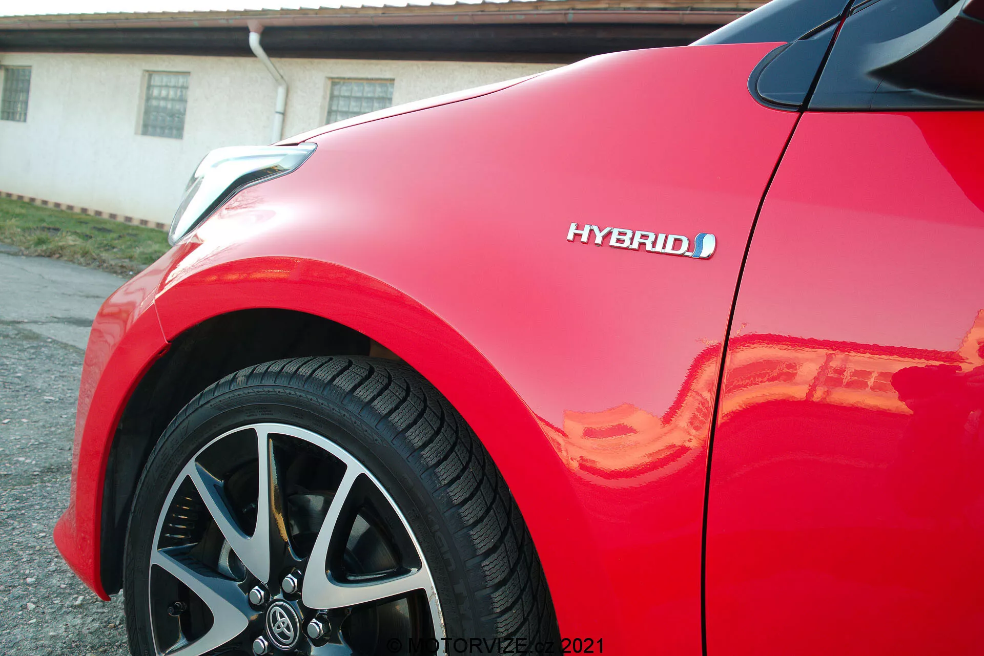 Vue rapprochée d'une Toyota Yaris 2020 rouge, se concentrant sur le panneau avant et la roue en alliage multi-branches. L'insigne hybride est visible, mettant en avant la technologie écologique du véhicule. La peinture brillante reflète la lumière du soleil, soulignant la finition de haute qualité et le design aérodynamique et moderne de la voiture.