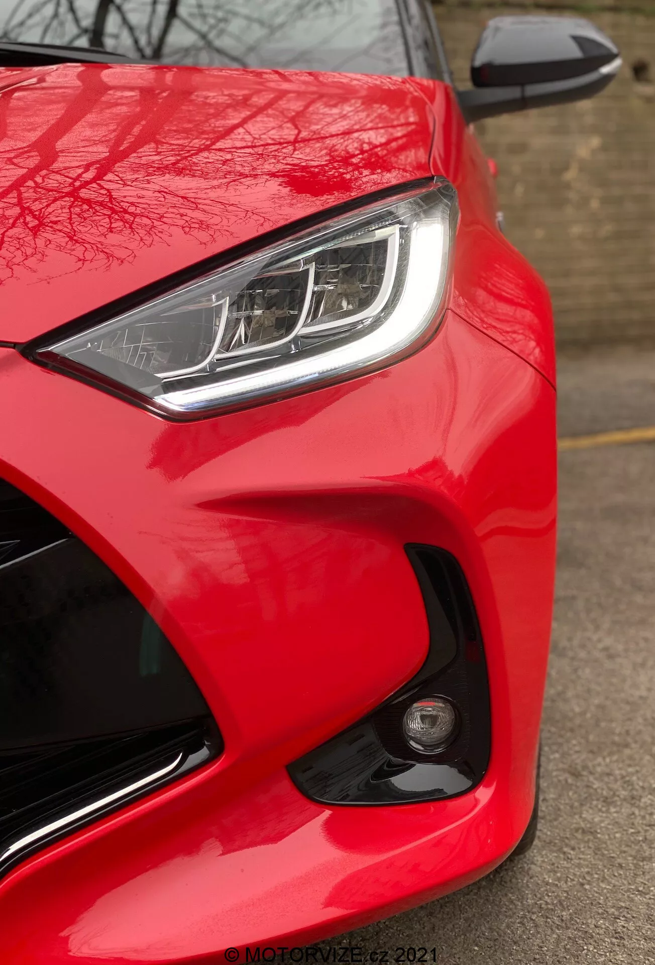 Vue rapprochée de la Toyota Yaris 2020, mettant en avant l'avant côté conducteur depuis un angle bas. L'image met en valeur la couleur rouge brillante et brillante de la voiture, le phare LED ou HID élégant avec des feux de jour distinctifs et le rétroviseur latéral partiellement visible de la même couleur que la carrosserie avec indicateur de clignotant intégré. Le design aérodynamique est souligné par les lignes sculptées et les courbes autour du phare et du capot. Un feu antibrouillard rond ou un capteur sur le pare-chocs avant suggère des fonctionnalités de sécurité avancées. L'absence de flou de mouvement indique que la voiture est stationnaire, et un filigrane 'MOTORZETZ. 2021' est visible en bas à droite.