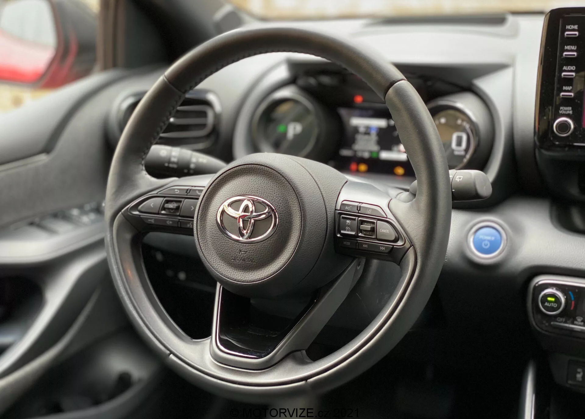 Vue intérieure d'une Toyota Yaris 2020, centrée sur le volant et le tableau de bord depuis la perspective du siège du conducteur. Le volant arbore le logo Toyota et des commandes visibles pour l'info-divertissement et les systèmes d'aide à la conduite. Derrière, on aperçoit des parties du tableau de bord numérique et de la console centrale avec les boutons de commande de la climatisation et les aérateurs.