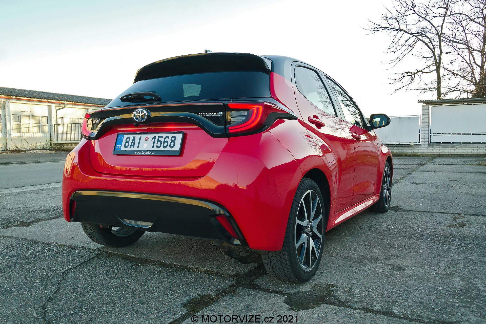 Voiture de tourisme Toyota Yaris 2020 rouge vif photographiée sous un angle trois quarts arrière, mettant en évidence les feux arrière à LED, le becquet arrière, l'emblème du modèle hybride, les jantes en alliage à plusieurs branches et les fonctionnalités d'assistance à la conduite visibles telles que les capteurs de stationnement et la caméra arrière, capturée depuis une perspective légèrement inférieure à la hauteur de la voiture.