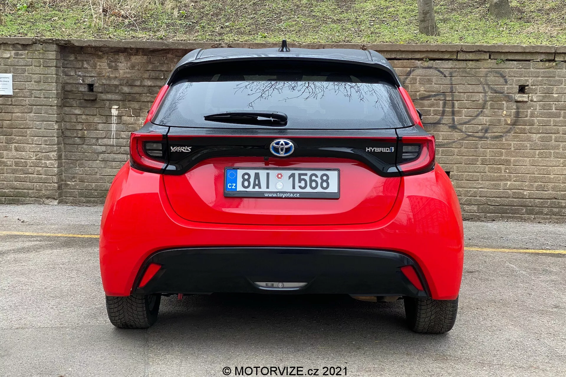 Vue arrière d'une Toyota Yaris Hybrid rouge de 2020, centrée et prise sous un angle direct. L'image révèle toute la largeur du véhicule, montrant l'emblème Toyota, les noms des modèles 'Yaris' et 'Hybrid', un aileron, une antenne montée sur le toit, une bande lumineuse de freinage haute et des feux arrière distinctifs. La lunette arrière avec un essuie-glace et une partie inférieure du pare-chocs pouvant potentiellement abriter des capteurs ou des réflecteurs sont également visibles. L'arrière-plan comprend un mur en briques ou une clôture et de la végétation, suggérant une voiture garée plus bas ou un angle de caméra plus bas.