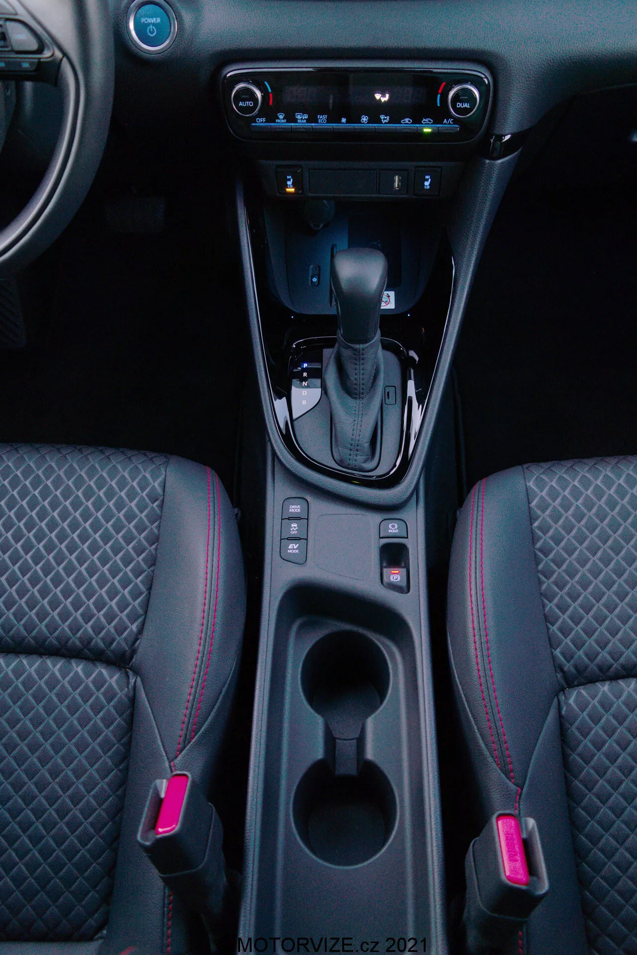 Vue verticale de l'intérieur d'une Toyota Yaris 2020 centrée sur la console centrale, mettant en vedette le levier de vitesse en position 'P', bouton de frein de stationnement électronique, deux porte-gobelets éclairés, espace de rangement, contrôle climatique numérique, système multimédia à écran tactile, port USB, bouches d'aération et sièges avant bicolores avec surpiqûres en motif diamant et surpiqûres rouges.