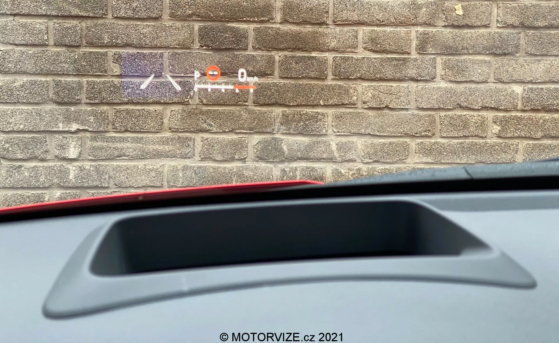 Vue rapprochée de l'intérieur d'une Toyota Yaris 2020 montrant une perspective partielle depuis le siège du conducteur vers l'arrière. L'accent est mis sur la tablette arrière avec un feu de freinage haut et un aileron arrière intégré. L'image montre également une partie du matériau intérieur tandis qu'un mur de briques à l'extérieur suggère que la voiture est garée près d'un bâtiment.