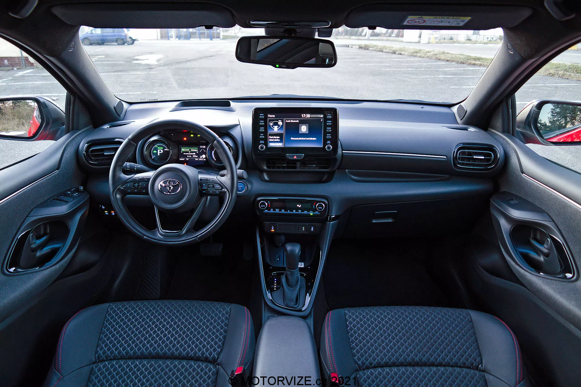 Vue intérieure d'une Toyota Yaris 2020 depuis la perspective du siège conducteur, présentant un tableau de bord moderne avec un volant équipé de commandes intégrées, un tableau de bord partiellement visible avec des écrans numériques et un système d'infotainment central avec interface tactile. Le tableau de bord comporte des aérations et des boutons de contrôle du climat, tandis que la console centrale possède des commandes pour les modes de conduite, un sélecteur de boîte automatique avec un soufflet en cuir, des porte-gobelets et un compartiment de rangement. Les sièges en tissu ont un motif design et les panneaux intérieurs des portes incluent des commandes pour les fenêtres électriques et les rétroviseurs.