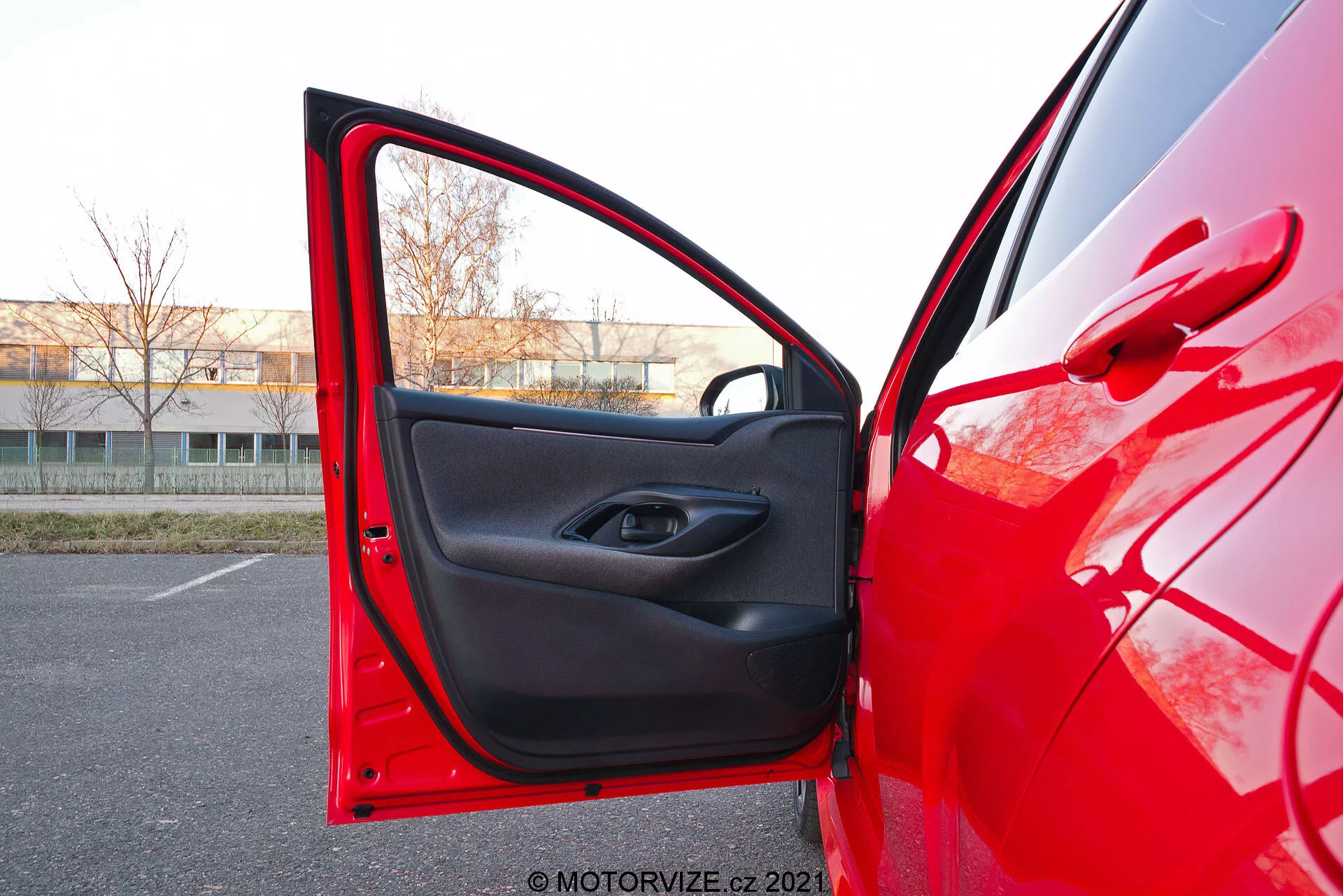 Vue en gros plan du côté conducteur d'une Toyota Yaris rouge de 2020 avec la porte avant ouverte, capturée sous un angle légèrement bas. L'appareil photo est positionné à l'extérieur, près de la porte, pointant vers l'intérieur, montrant la peinture extérieure rouge brillante reflétant la lumière et la garniture intérieure noire au design minimaliste. La porte possède un bouton de commande de fenêtre électrique dans l'indentation de la poignée et une poche de rangement en dessous.