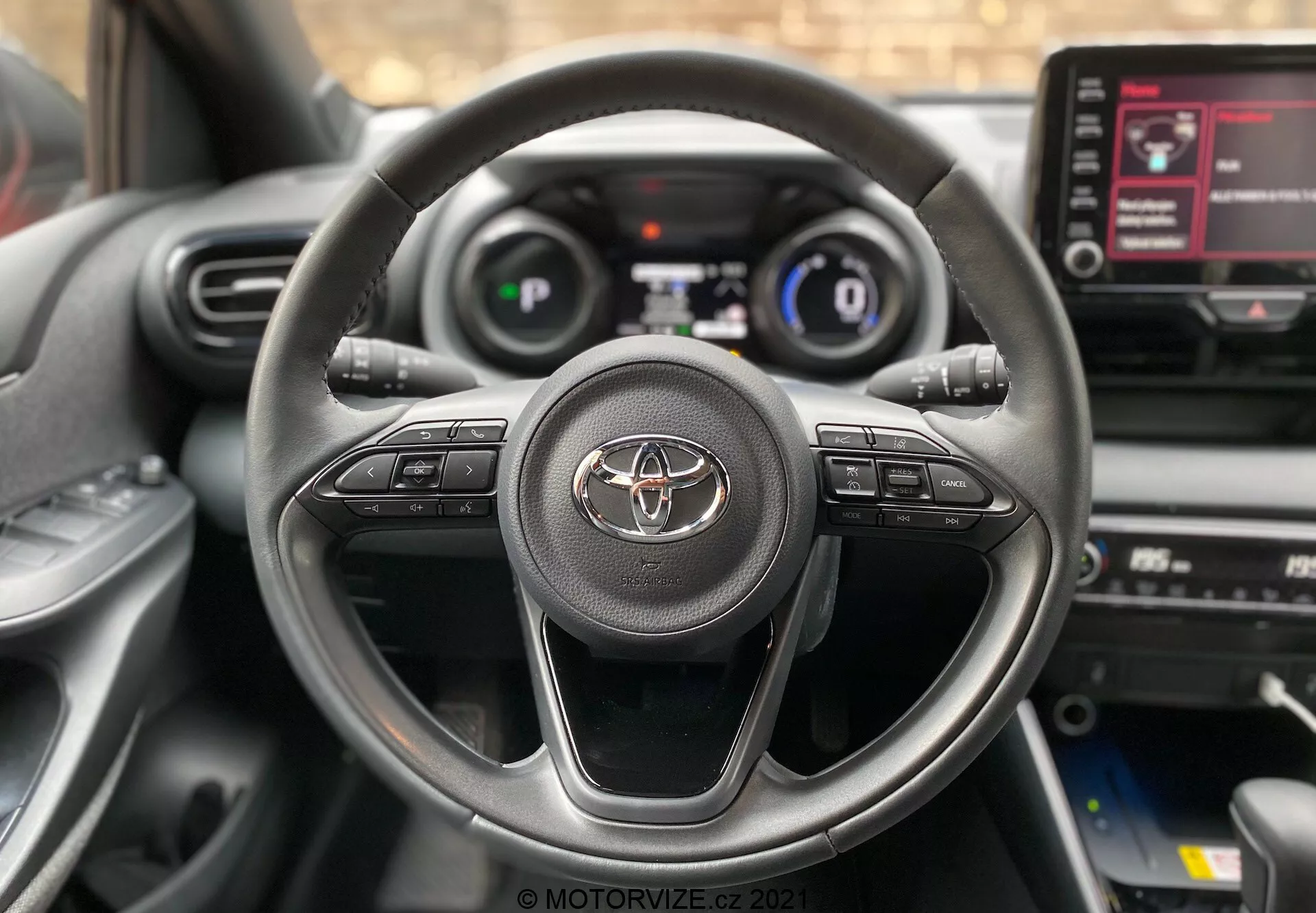 Gros plan de l'intérieur de la Toyota Yaris 2020 depuis le siège du conducteur, avec un volant trois branches portant l'emblème Toyota et des boutons de commande pour l'audio, les appels mains libres et le régulateur de vitesse. Le tableau de bord numérique affichant la vitesse et les informations de conduite essentielles est partiellement visible. Console centrale avec écran tactile info-divertissement affichant les options de menu également visible.