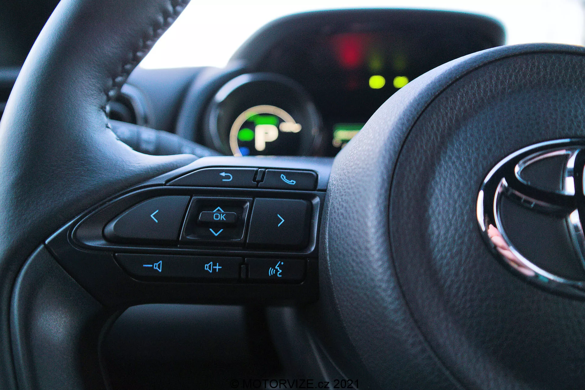 Vue intérieure rapprochée d'une Toyota Yaris 2020 depuis la perspective du siège du conducteur, mettant en avant le volant avec des commandes multifonctions et le tableau de bord en arrière-plan. Le volant domine les deux tiers inférieurs de l'image, avec des flèches, des commandes vocales, de volume et du système de divertissement visibles. Le tableau de bord partiellement visible est illuminé avec un affichage numérique vert et est flou en raison d'une faible profondeur de champ. La lumière naturelle illumine l'intérieur, soulignant le confort et une interface conviviale.