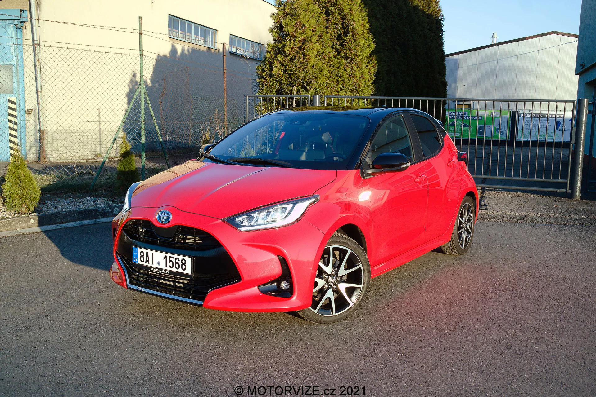 Image d'une Toyota Yaris 2020 hatchback rouge vif garée en angle, montrant à la fois les profils avant et latéral. Le design des phares est aiguisé et angulaire, la calandre présente un motif en nid d'abeille avec un emblème central, et le pare-chocs avant affiche des contours visibles avec des feux antibrouillard. Le profil latéral révèle des jantes en alliage bicolore, des poignées de porte et des rétroviseurs de couleur assortie, avec des lignes nettes et des contours tout au long du côté. La peinture brillante suggère un état neuf ou bien entretenu. L'arrière-plan comprend un ciel clair, des structures industrielles et des ombres douces, indiquant un cadre extérieur, probablement une zone de stationnement près d'une installation commerciale ou industrielle.