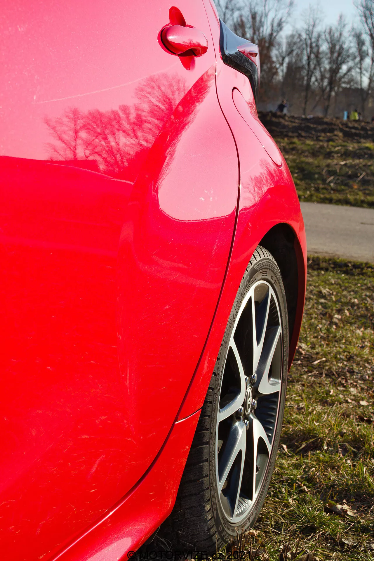 Vue latérale rapprochée du panneau arrière d'une Toyota Yaris 2020 rouge prise avec un angle légèrement ascendant mettant en avant la finition rouge vif, la jante en alliage à rayons multiples, le motif de la bande de roulement du pneu, la courbure propre du panneau arrière et du pare-chocs, le reflet brillant de la peinture avec des arbres et un ciel bleu clair, et le bouchon de carburant et le détail de garniture visibles.
