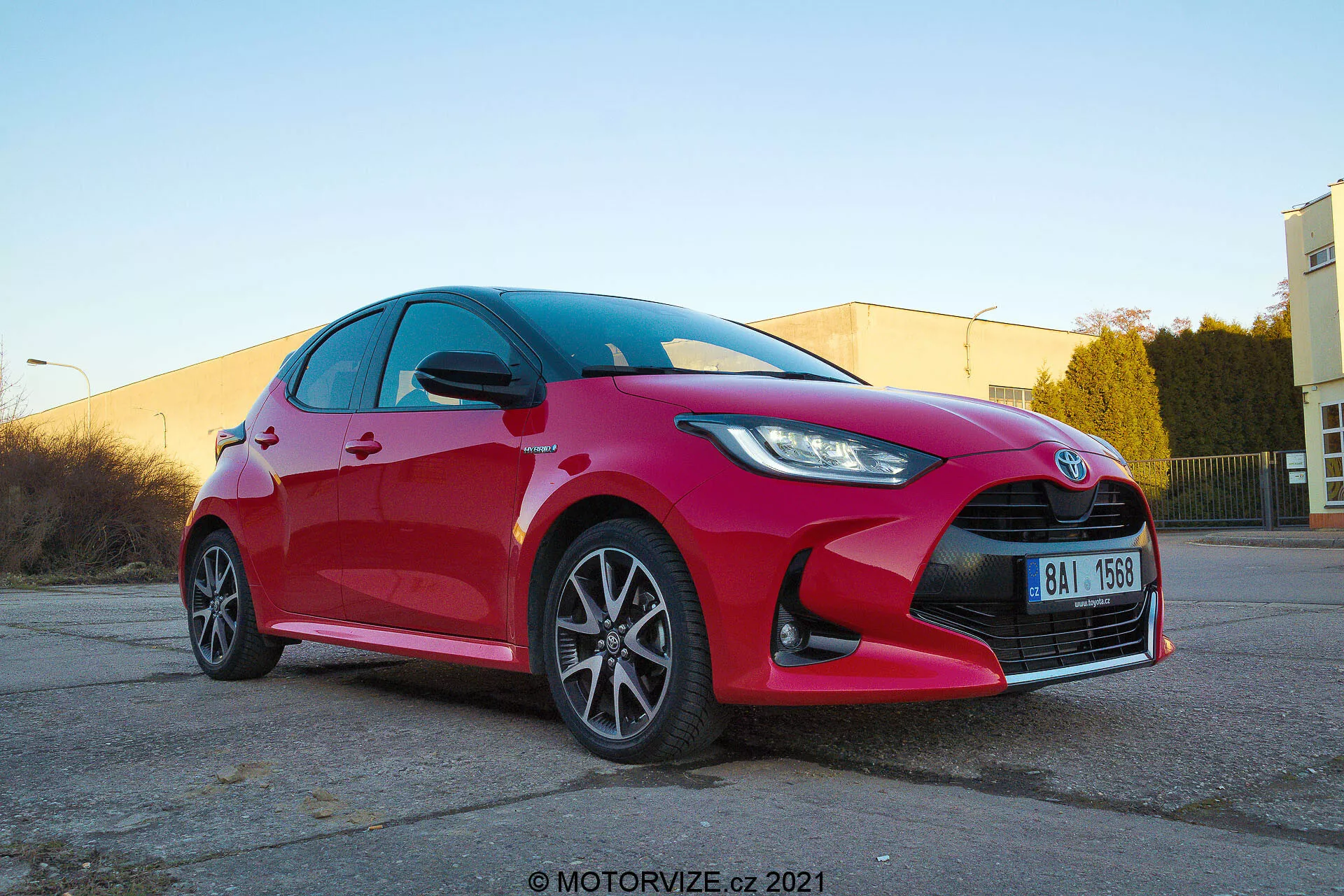Vue de trois quarts avant en contre-plongée d'une Toyota Yaris hatchback rouge vif de 2020 montrant des phares LED ou HID angulaires et nets, une calandre proéminente avec emblème central, des roues en alliage multi-rayons, des clignotants intégrés dans les rétroviseurs latéraux, des surfaces réfléchissantes propres et un possible système de freinage orienté performance. Capturé dans un parking industriel avec un éclairage chaleureux et filigrane motorvize.cz 2021.