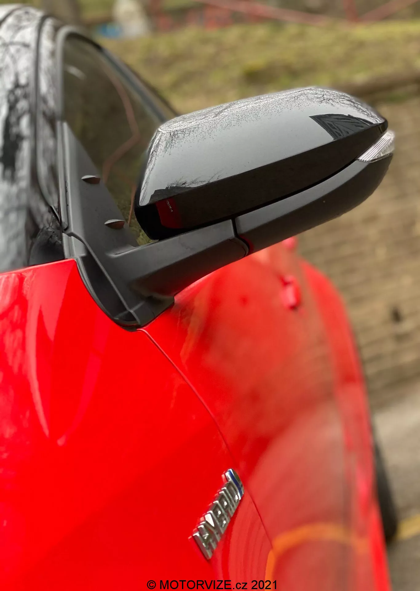 Gros plan sur le rétroviseur latéral déployé d'une Toyota Yaris 2020, avec un boîtier noir aérodynamique élégant contrastant avec la carrosserie rouge et brillante de la voiture. Le rétroviseur met en avant des touches technologiques possibles comme le réglage électrique, le chauffage et les clignotants intégrés, bien que les caractéristiques spécifiques ne soient pas confirmées. Les gouttes d'eau suggèrent une pluie récente ou un lavage de voiture, tandis qu'un arrière-plan flou et une faible profondeur de champ maintiennent l'attention sur le rétroviseur. Une partie du badge sur l'aile avant est visible mais floue.