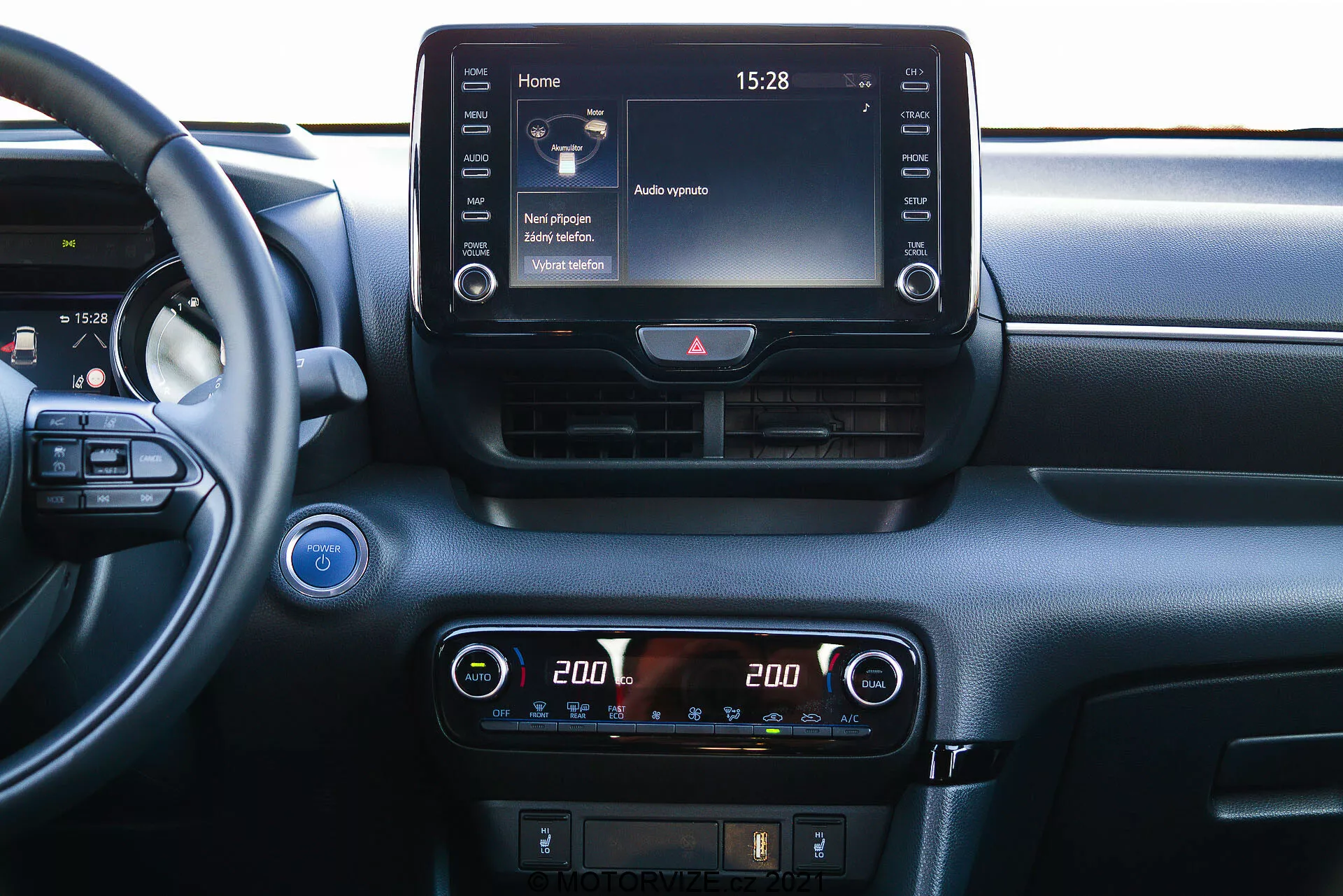 Vue rapprochée du tableau de bord central d'une Toyota Yaris 2020 depuis une perspective intérieure. Le volant est partiellement visible sur la gauche, suggérant un angle de vue depuis le siège passager ou central. Le tableau de bord comporte un écran tactile d'infodivertissement proéminent en haut de la console centrale, affichant un écran d'accueil avec diverses icônes et l'heure '15:28'. En dessous, un affichage numérique du contrôle du climat avec des réglages de température à double zone, chaque côté réglé sur '20.0' degrés Celsius. Les commandes supplémentaires pour le dégivrage et peut-être l'assistance au conducteur sont visibles sous les commandes du climat. Au-dessus de l'écran d'infodivertissement, il y a deux bouches d'aération horizontales avec un bouton de feux de détresse entre elles. Un bouton de démarrage/arrêt est partiellement visible à l'extrême gauche, indiquant une technologie de démarrage sans clé. Une grille de haut-parleur est située dans le coin supérieur gauche. Le matériau du tableau de bord présente des éléments à la fois lisses et texturés, suggérant une attention au confort et à la technologie.