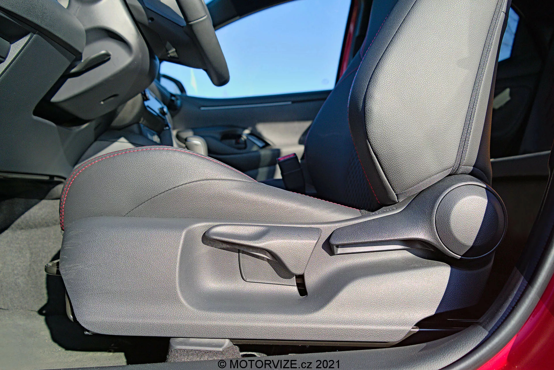 Vue intérieure détaillée de la Toyota Yaris 2020 se concentrant sur la porte et le siège conducteur, mettant en avant la sellerie bicolore, les surpiqûres décoratives, le design profilé pour un soutien ergonomique, les bourrelets latéraux réglables manuellement et un levier de réglage manuel bien en vue. Le cadre de la porte est partiellement visible, suggérant que la porte est ouverte.