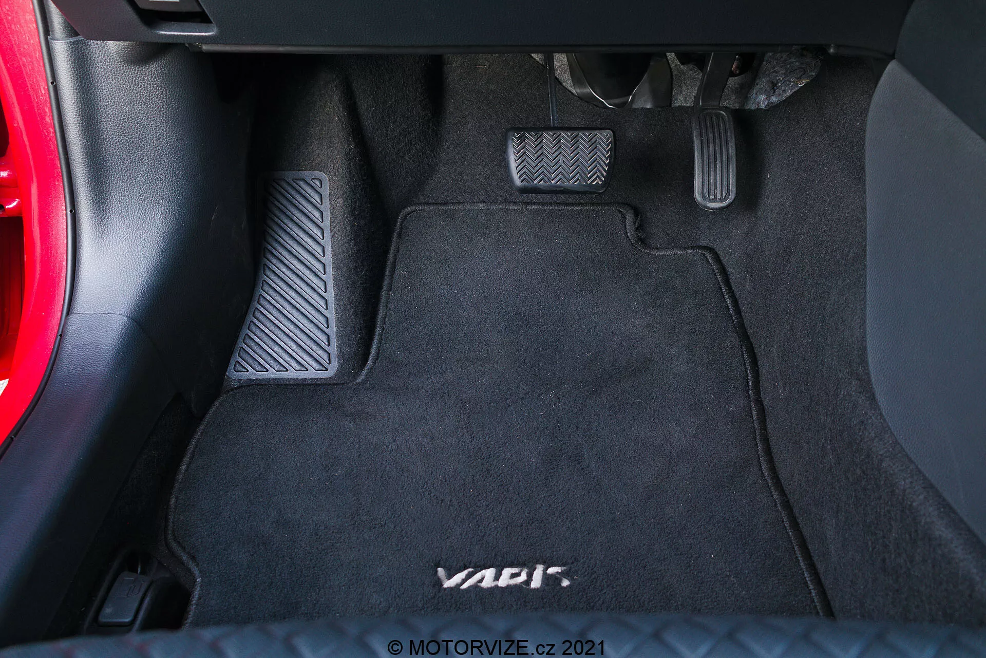 Vue rapprochée de l'espace pour les pieds côté conducteur d'un véhicule, mettant en avant trois pédales en finition métallique avec inserts en caoutchouc pour une configuration à transmission manuelle sur un tapis de sol noir marqué 'VAG'. La pédale de gauche est l'embrayage, celle du milieu est le frein et celle de droite est l'accélérateur. Le bas du tableau de bord et une section de la porte de la voiture sont partiellement visibles.