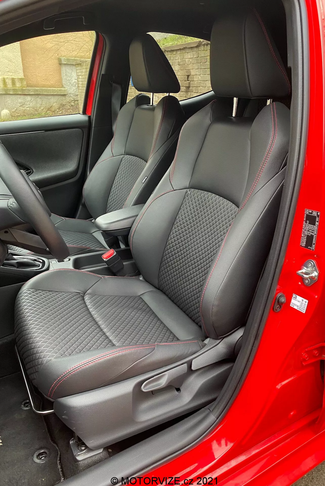 Intérieur de la Toyota Yaris 2020 vu du côté passager avec la porte ouverte, mettant en évidence des sièges en tissu texturé de qualité supérieure avec des motifs à losanges et des accents rouges, des appuie-têtes réglables, des commandes de console centrale, et un panneau de porte texturé en fibre de carbone avec des commandes électriques des fenêtres.