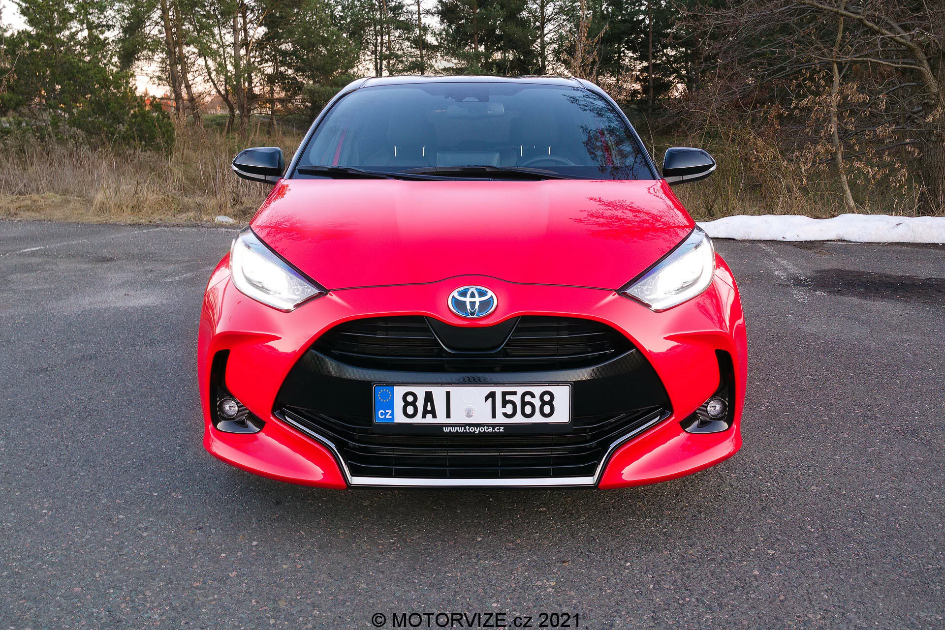 L'image montre une vue de face d'une Toyota Yaris 2020 rouge, prise sous un léger angle, se concentrant sur la calandre avant, les phares et le capot, avec la plaque d'immatriculation entièrement visible. La voiture présente des éléments de design distinctifs, notamment l'emblème Toyota centré sur la calandre, des phares angulaires nets se rétrécissant vers la calandre et des courbes complexes sur le capot, mettant en valeur son design aérodynamique et sportif. La photo, capturée pendant la journée avant le crépuscule, suggère que la voiture est garée sur une surface plane dans un environnement qui pourrait être la fin de l'automne ou le début du printemps.