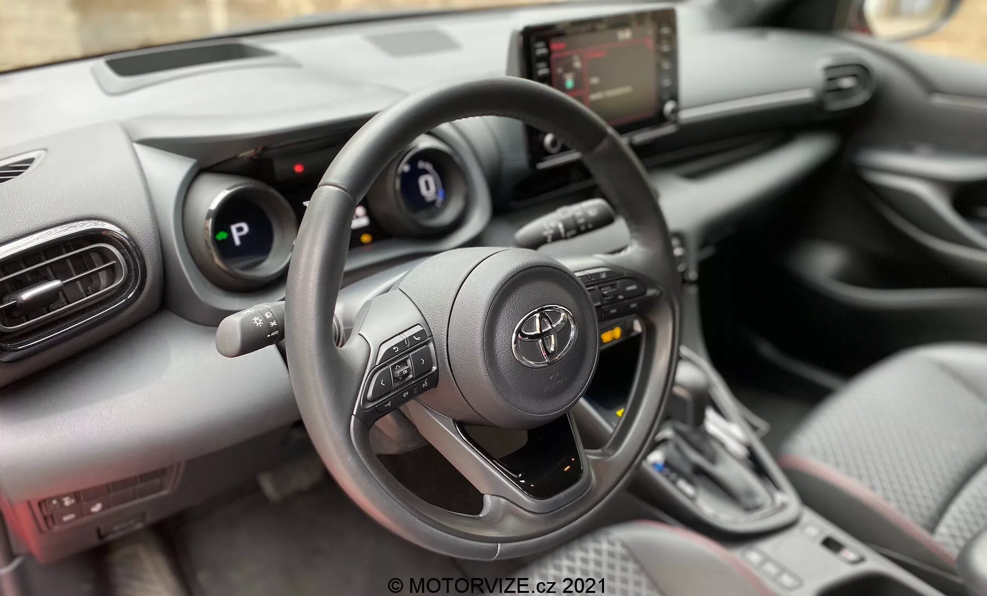 Cette image présente l'intérieur d'une voiture, en se concentrant sur la perspective du conducteur. La prise de vue est un gros plan, vue intérieure de face, avec l'angle de la caméra positionné pour capturer principalement le volant, le tableau de bord et la console centrale. La photographie est probablement prise depuis le siège du conducteur ou peut-être juste au-dessus, offrant un point de vue similaire à celui d'une personne assise derrière le volant. Le volant est proéminent au premier plan, centré dans le cadre, et occupe une partie substantielle de l'image. Il s'agit d'un design à trois branches avec des commandes multifonctions montées sur les branches gauche et droite. Derrière le volant, le tableau de bord comporte un affichage numérique encadré par des jauges analogiques. À droite du volant, la console centrale comprend un écran tactile infotainment, un panneau de contrôle de la climatisation et un sélecteur de vitesse, indiquant un véhicule moderne avec des fonctions interactives.
