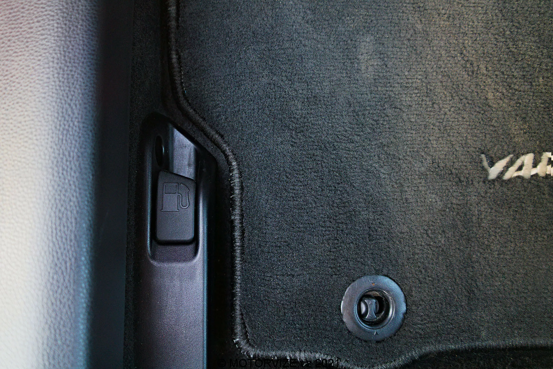 Vue en gros plan de l'intérieur de la Toyota Yaris 2020, capturant le côté inférieur gauche près de la porte. Sont visibles un tapis de sol foncé avec un texte brodé partiellement visible, un crochet de retenue circulaire en métal, le seuil de porte en plastique avec une garniture de protection, un levier de déverrouillage du coffre suggérant le côté conducteur et un schéma de couleurs bicolore avec textures contrastantes de tapis et de plastique lisse.