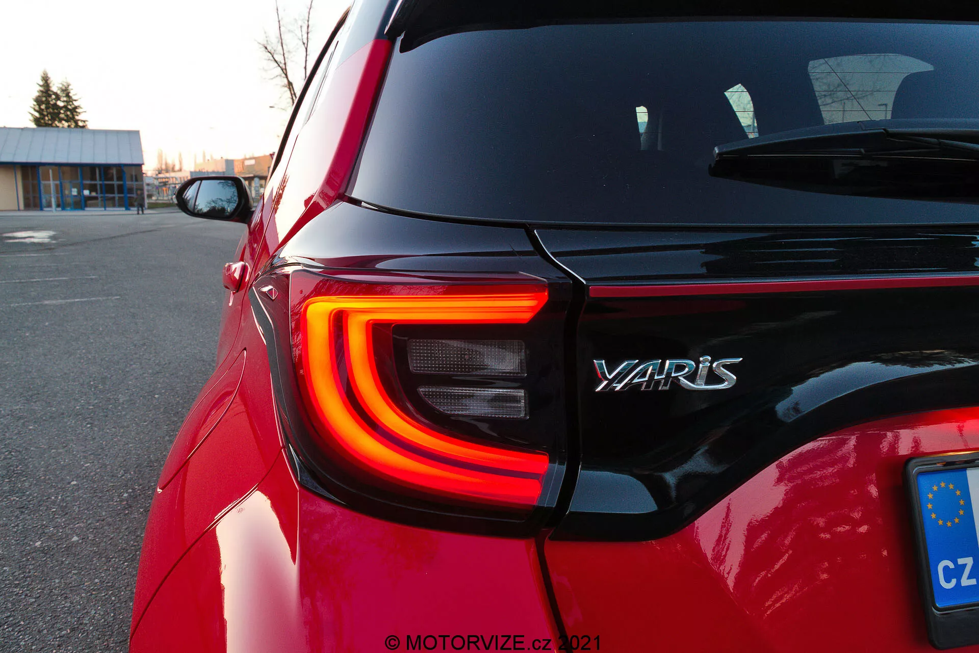 Gros plan sur la section arrière d'une Toyota Yaris rouge de 2020, montrant le feu arrière gauche à DEL avec sa forme en C distinctive, le nom du modèle 'YARIS' sur le hayon arrière et une plaque d'immatriculation de l'Union Européenne de la République Tchèque. La prise de vue en angle oblique met en évidence les contours et la réflectivité de la peinture de la voiture au crépuscule sur un parking, avec des arrière-plans flous qui attirent l'attention sur les détails du design.