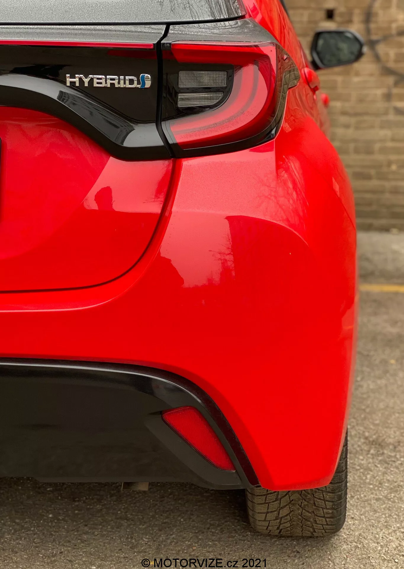 Vue arrière inclinée d'une Toyota Yaris hybride rouge 2020, en se concentrant sur le quart arrière côté droit. Capture des fonctionnalités telles que le badge 'HYBRID', le feu arrière LED moderne, le léger pli aérodynamique du pare-chocs arrière et un capteur de stationnement visible. La peinture brillante et le dessin de bande de roulement bien entretenu indiquent un véhicule neuf ou bien entretenu.