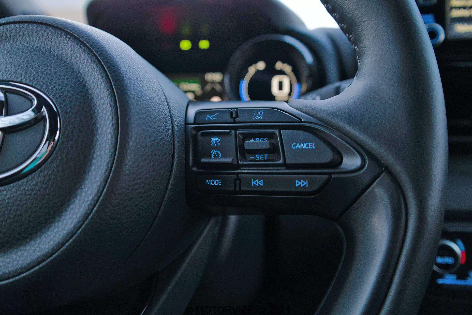 Photo de l'intérieur d'une Toyota Yaris 2020 depuis le siège du conducteur, focalisée sur le côté gauche du volant multifonction. Les boutons tactiles, y compris l'icône de la fonction de sécurité, le régulateur de vitesse avec étiquette 'SET', le bouton d'annulation, 'MODE' pour les multimédias et les commandes de volume '+', '-', sont bien visibles. Le véhicule est en mode 'P' parking, avec le tableau de bord et les éléments en arrière-plan légèrement flous.