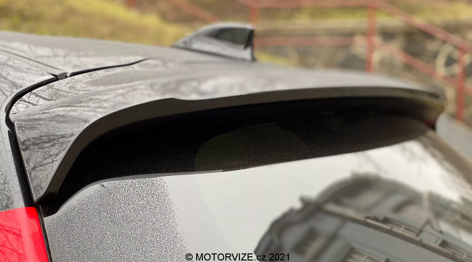 Gros plan sur le spoiler arrière de la Toyota Yaris 2020 avec une peinture noire contrastant avec la carrosserie métallique. L'image capture des gouttelettes d'eau sur le spoiler, suggérant des conditions humides, et un profil latéral révélant son design légèrement surélevé pour une meilleure aérodynamique. L'arrière-plan flou en extérieur ajoute du contexte à la finition premium de la voiture et le feu arrière LED visible suggère des fonctionnalités modernes.