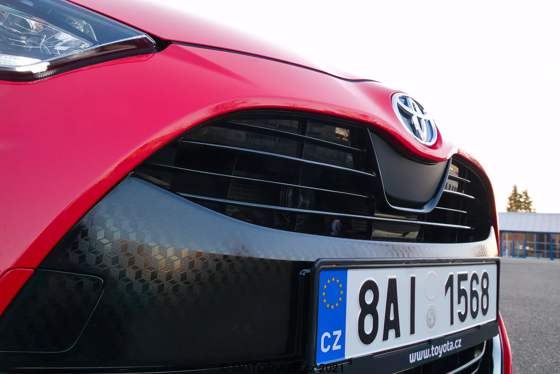 Image en gros plan d'une Toyota Yaris 2020 rouge se concentrant sur le côté avant gauche, montrant le capot, le phare moderne avec une technologie LED ou HID potentielle, la calandre avec l'emblème Toyota et un motif en nid d'abeille, la peinture rouge brillante et le pare-chocs avant partiellement visible avec une prise d'air inférieure. La plaque d'immatriculation de l'Union Européenne indique une immatriculation en République Tchèque.