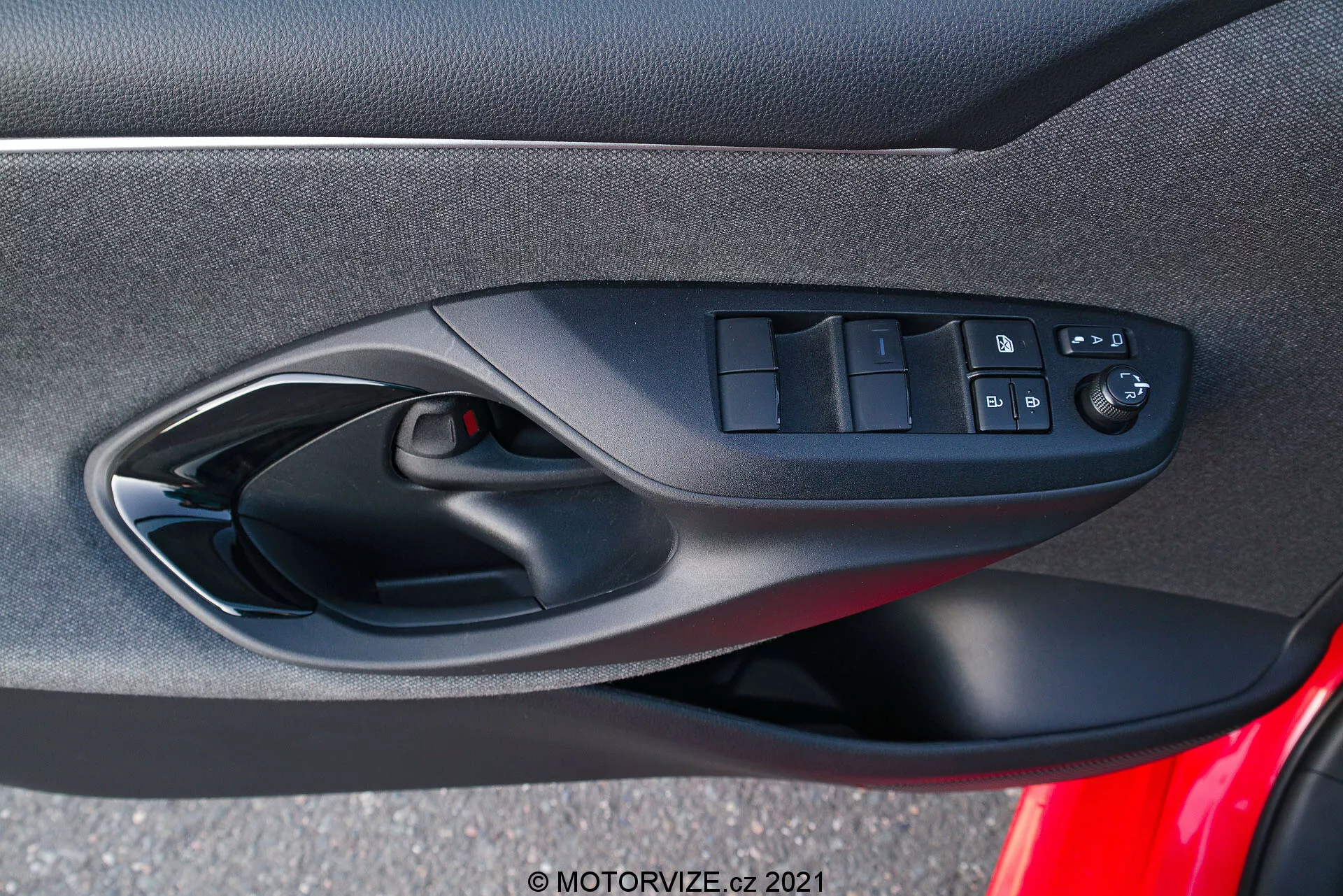 Panneau de porte intérieur d'une Toyota Yaris 2020 se concentrant sur les commandes de la porte côté conducteur conçues de manière ergonomique, y compris les interrupteurs des fenêtres électriques, le bouton de verrouillage des portes et les commandes réglables électriquement des rétroviseurs, capturé depuis la perspective d'un occupant à l'intérieur du véhicule. La photo met en évidence le mélange de matériaux en tissu ou composite doux, le schéma de couleur noir et une grille de haut-parleur visible faisant partie du système audio de la voiture.