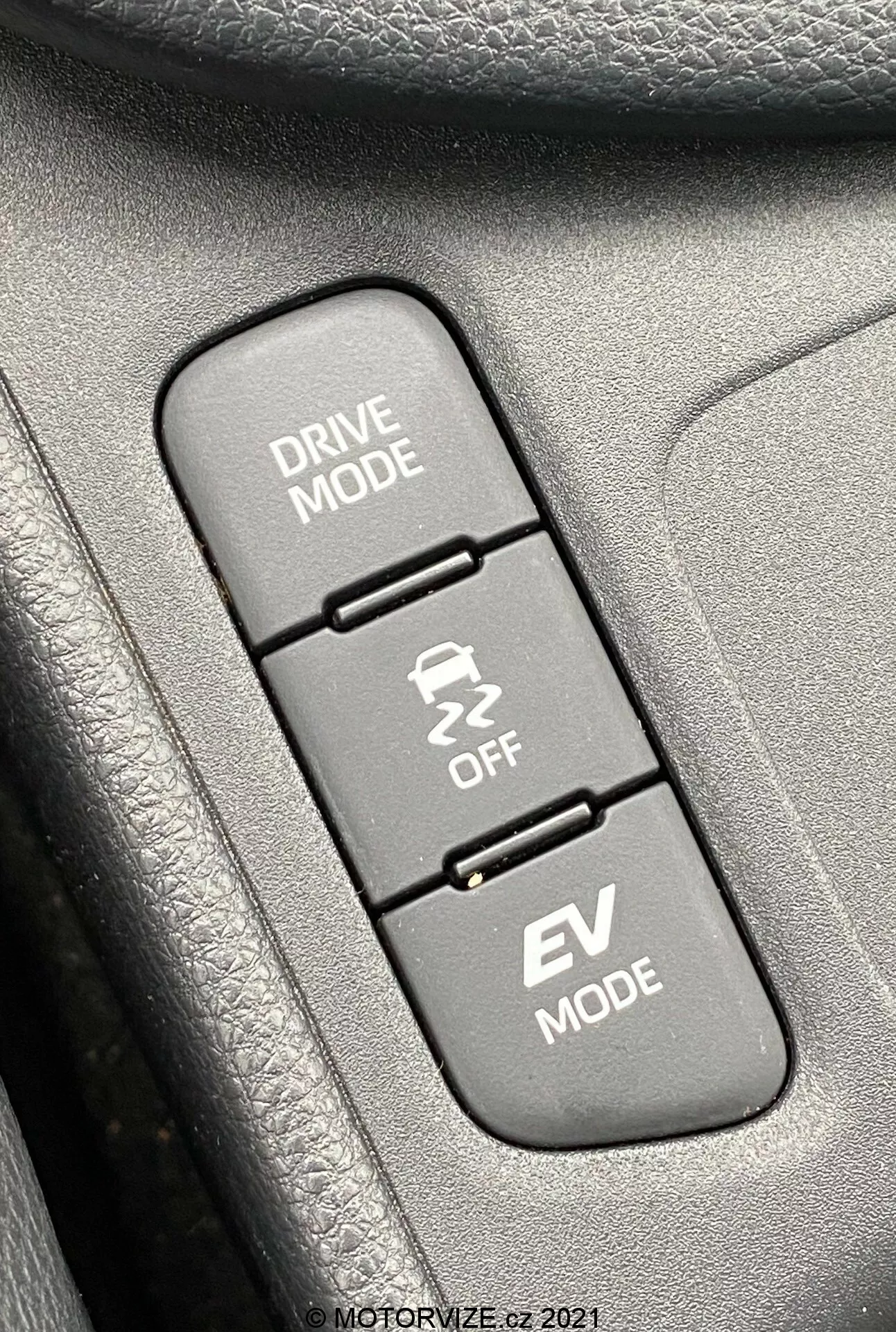Gros plan de l'intérieur d'une Toyota Yaris 2020, se concentrant sur trois boutons de contrôle alignés verticalement liés aux modes de conduite. De haut en bas : 'DRIVE MODE' avec une icône de route sinueuse, 'Traction Control OFF' avec une icône de voiture dérapant et 'EV MODE' indiquant la fonction de véhicule électrique. Les boutons sont montés sur une surface texturée, probablement une partie du panneau de porte ou de la console centrale. L'image est nette sur les boutons, tandis que les textures environnantes sont légèrement floues, éclairée uniformément par la lumière du jour pour une visibilité claire.