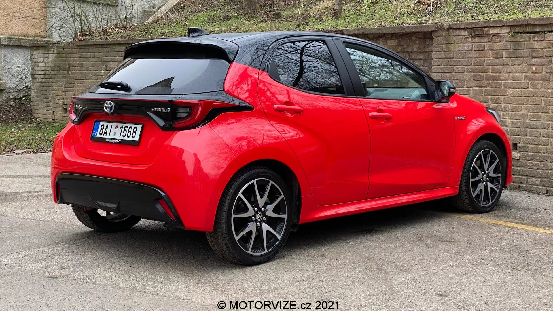 Vue arrière trois quarts d'une voiture hatchback moderne rouge vif, avec des lignes nettes et des plis, un becquet de toit, des feux arrière à LED, une antenne aileron de requin, des roues en alliage à rayons multiples et placée sur un parking avec une peinture réfléchissante et bien entretenue, suggérant un modèle récent ou bien entretenu dans une version sportive.
