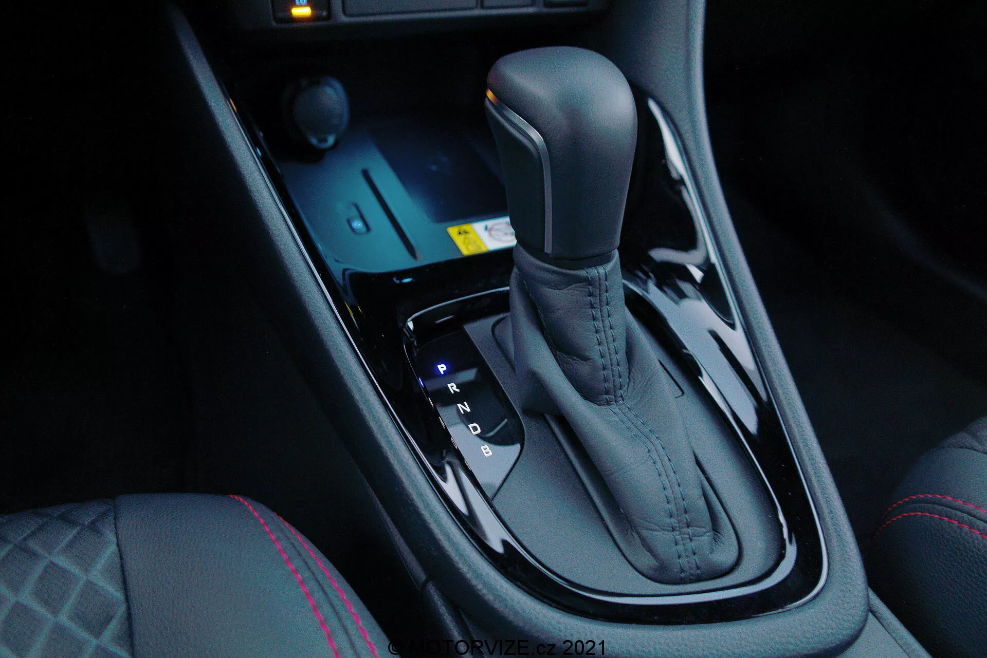 Vue rapprochée du levier de vitesses automatique de la Toyota Yaris 2020, vue depuis le côté passager avant avec un angle vers le bas, mettant en avant la finition métallique élégante et le design ergonomique. Le levier de vitesses est positionné au centre entre les sièges conducteur et passager avant et comprend les positions Park, Reverse, Neutral et Drive, ainsi que des options de changement manuel (+ et -). Devant le levier de vitesses se trouvent des commandes électroniques pour les modes de conduite et le contrôle de la traction, ainsi qu'un bouton de démarrage éclairé en bleu. La console centrale sombre et brillante ajoute une touche de luxe moderne, complétée par les contrôles de chauffage des sièges et les coutures distinctives sur le siège du conducteur, indiquant un confort accru et des dynamiques de conduite avancées.