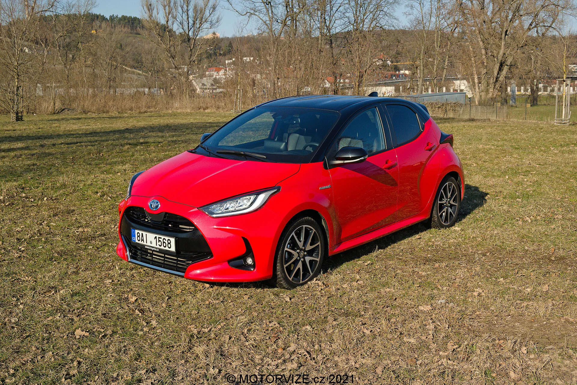 L'image montre une Toyota Yaris 2020 rouge éclatante, reposant sur un terrain inégal et herbeux dans une zone semi-rurale, vue en perspective trois quarts. Le design moderne et aérodynamique de la voiture présente des angles vifs autour des phares et une apparence compacte et sportive. Des roues à plusieurs rayons foncés et des phares LED stylisés sont visibles. Les vitres latérales et le pare-brise avant offrent un aperçu sombre de l'intérieur, ce qui laisse entrevoir d'éventuels systèmes automobiles avancés.