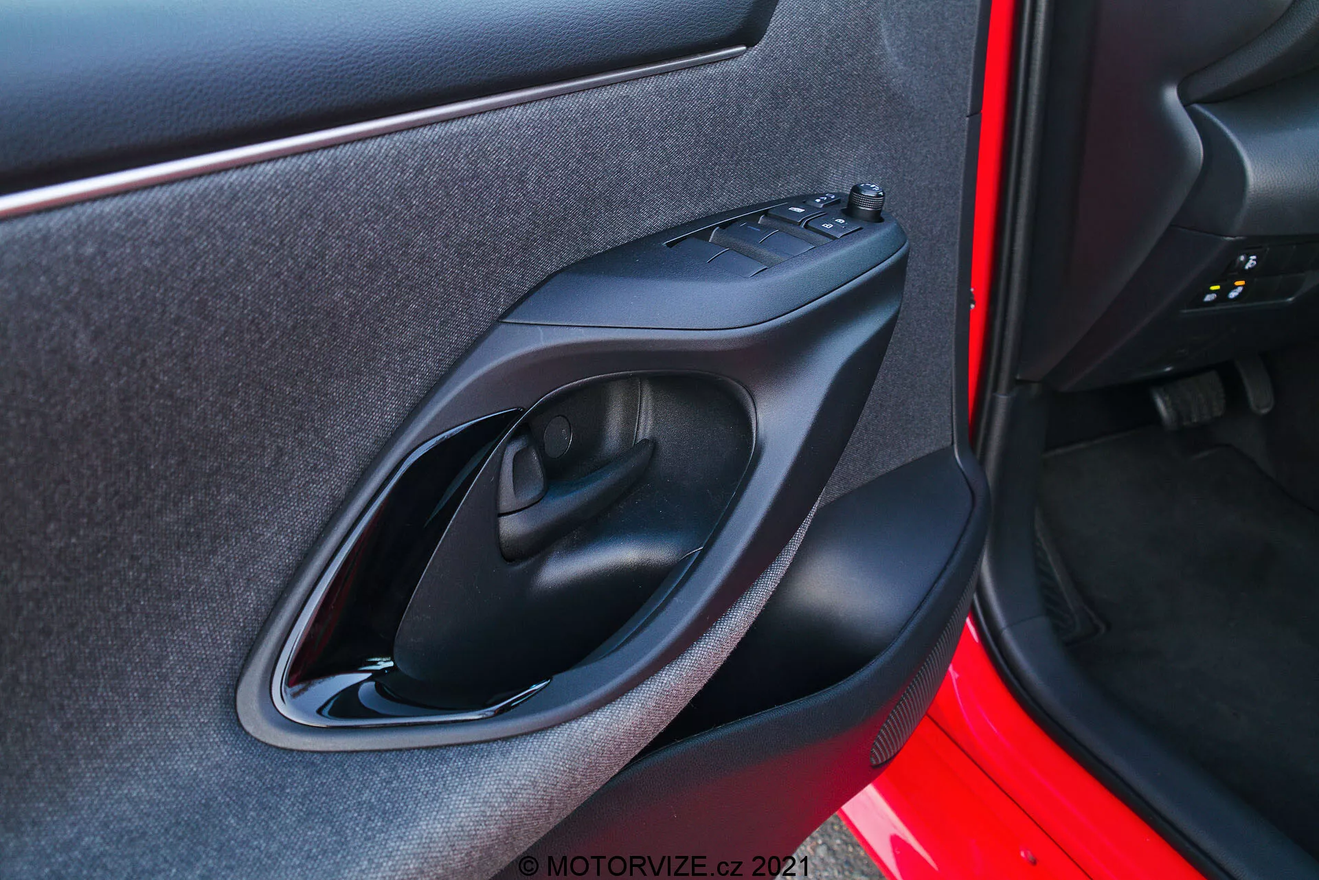 Gros plan du panneau de porte intérieur d'une Toyota Yaris 2020 pris sous une perspective extérieure. L'image se concentre sur le panneau de porte côté droit avec des caractéristiques de conception et de technologie telles qu'une poignée de porte en plastique noir mat, un panneau de contrôle des vitres électriques, un accoudoir ergonomique contourné et une petite poche de rangement. L'extérieur de la porte est peint en rouge vif.