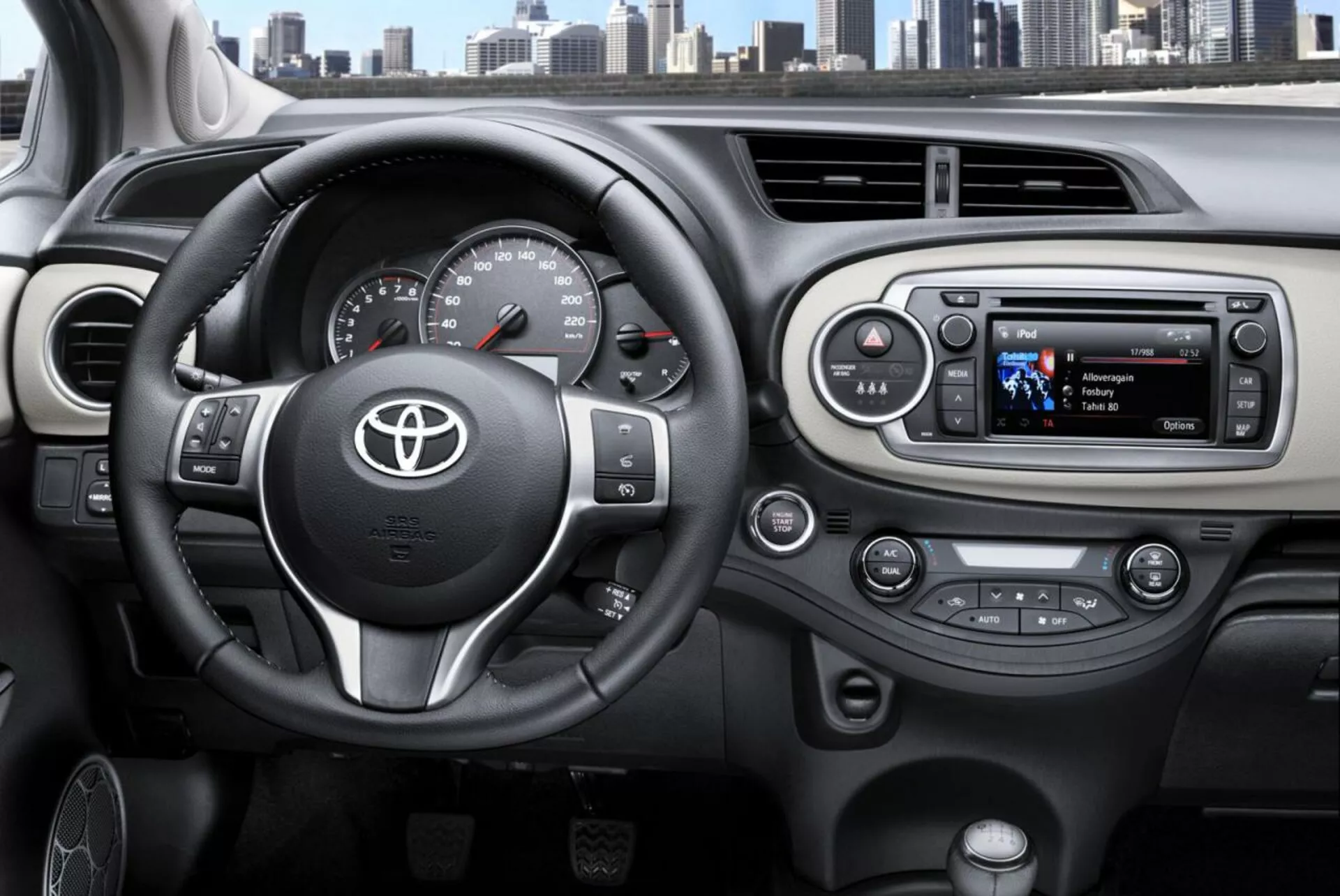 Vue intérieure du Toyota Yaris 2014 du point de vue du conducteur mettant en lumière le volant à trois branches avec emblème Toyota, commandes montées pour l'audio et téléphone mains-libres, compteur de vitesse et tachymètre analogiques avec un affichage numérique, et un système d'infodivertissement moderne avec boutons de contrôle du climat sur le tableau de bord