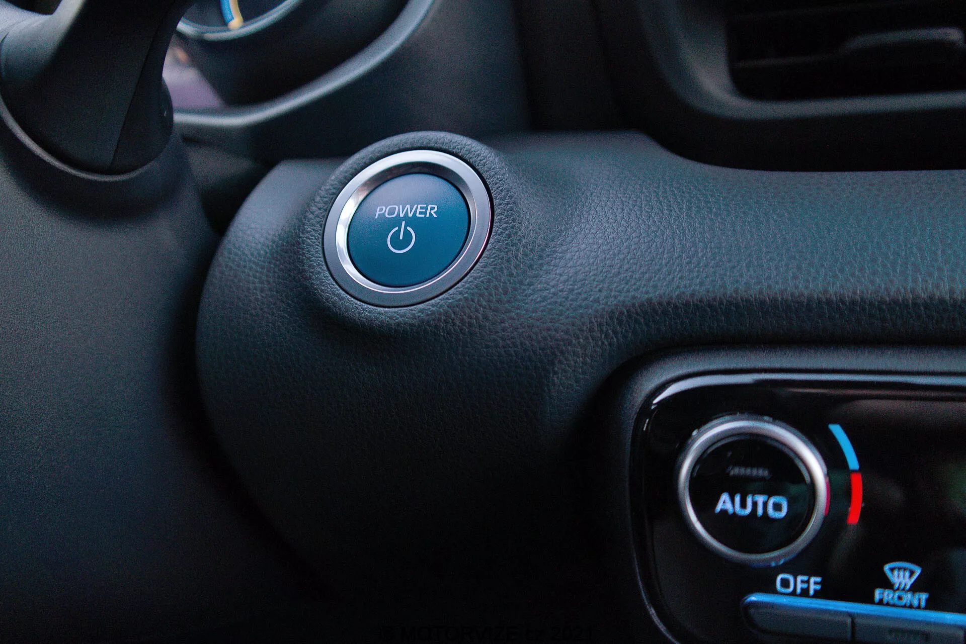 Vue intérieure du côté conducteur de la Toyota Yaris 2020 mettant en avant le bouton Start/Stop et le contrôle climatique. Le bouton de démarrage avec un symbole illuminé en bleu est proéminent dans le coin supérieur gauche, avec un accent argenté. En bas à droite, les cadrans de commande de la climatisation montrent une lumière bleue et une icône de flocon de neige pour la température froide, avec les étiquettes 'AUTO', 'OFF' et 'FRONT'. Un mélange de matériaux synthétiques noirs texturés et lisses dénote un style minimaliste avec des fonctionnalités de démarrage sans clé et de contrôle climatique automatique.