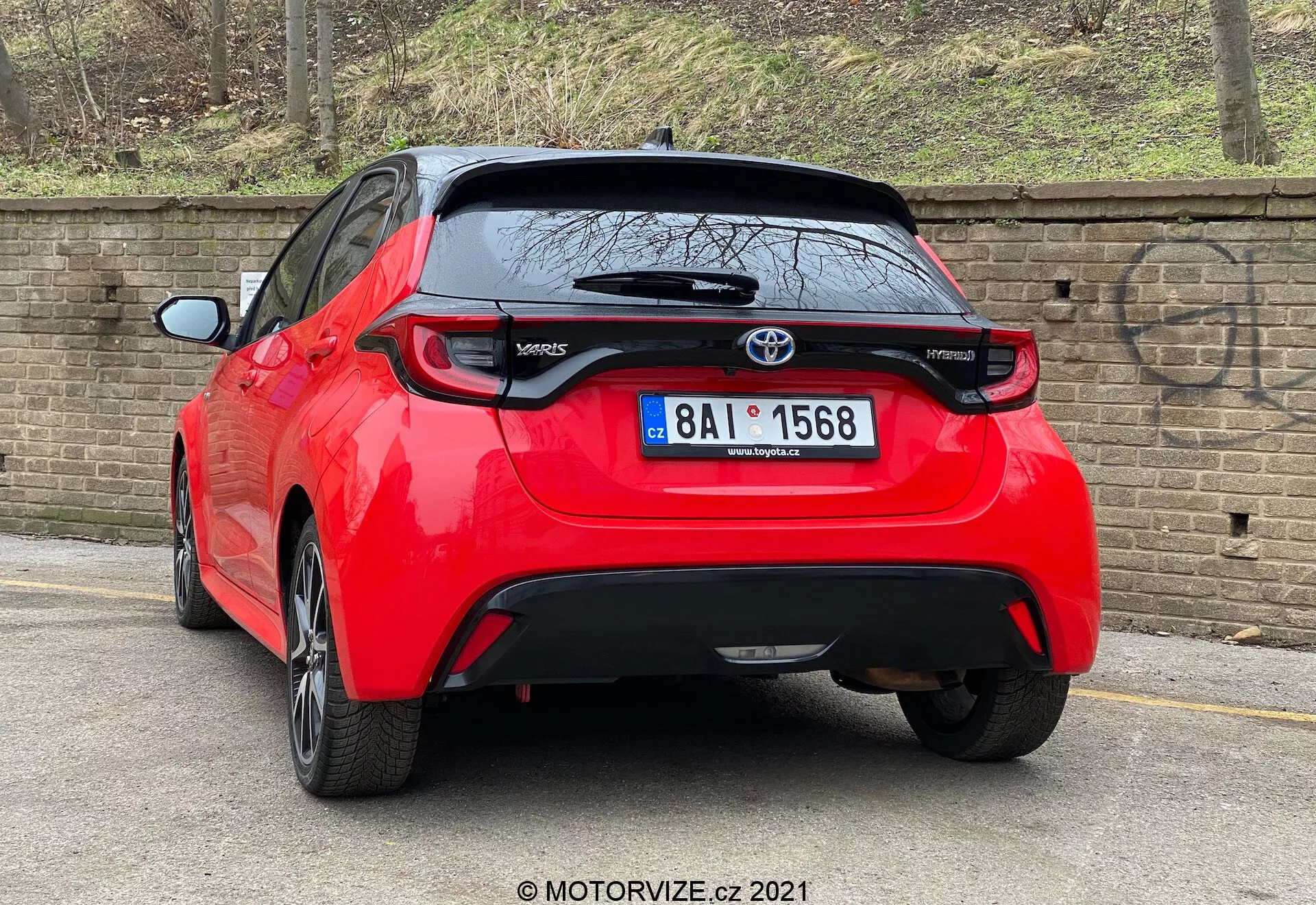 Une image haute résolution d'une Toyota Yaris 2020 rouge hatchback vue sous un angle arrière trois-quarts. Le véhicule est équipé de feux arrière distinctifs, d'un emblème Toyota central au-dessus d'une probable caméra de recul et du badge du modèle 'Yaris' en bas à gauche du hayon. Sous la zone de la plaque d'immatriculation se trouve une pièce de garniture noire qui pourrait être en plastique ou en fibre de carbone, avec un petit becquet arrière qui s'étend au-dessus de la lunette arrière. La voiture, garée près d'un trottoir sur une route pavée à côté d'un mur de briques, suggère un environnement urbain ou résidentiel.
