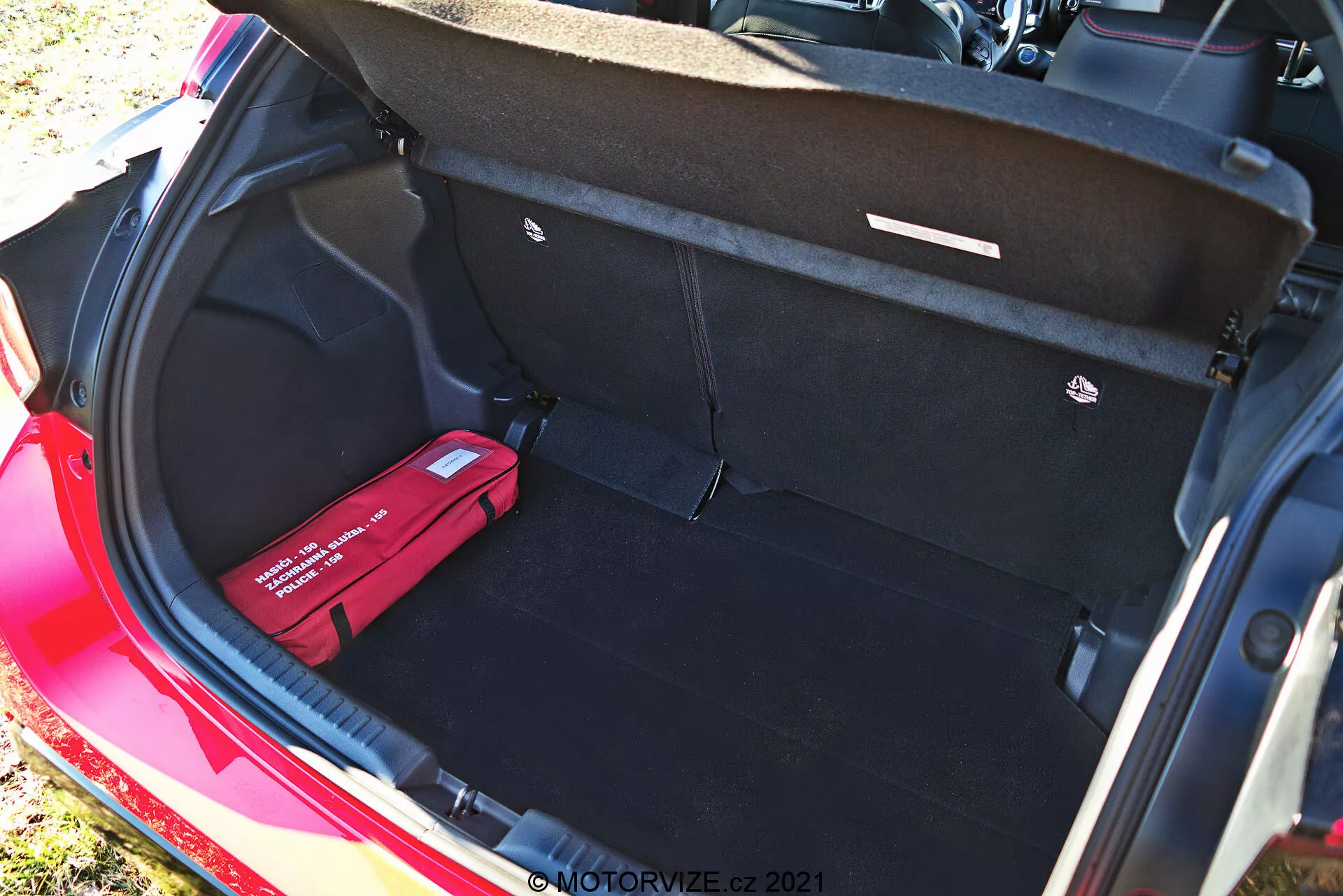 Coffre d'une Toyota Yaris 2020 rouge vu depuis un angle élevé, montrant son intérieur propre et spacieux avec un tapis noir, un couvercle de coffre ouvert avec des poignées de traction, un plancher de chargement plat, un crochet de chargement à droite et une trousse de premiers soins ou d'urgence rouge à gauche. Cette photographie prise en journée met en évidence la grande capacité de stockage et les fonctionnalités organisationnelles du véhicule.