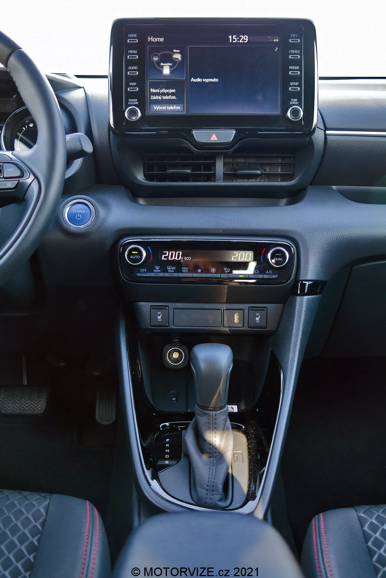 Vue intérieure d'une Toyota Yaris 2020 se concentrant sur la console centrale et les éléments du tableau de bord. La photo, prise depuis le siège passager avant, montre un système d'infodivertissement à écran tactile numérique affichant un écran d'accueil avec l'heure 15:29 et diverses options de menu, des aérateurs centraux et des commandes conviviales pour la climatisation et le chauffage avec un affichage numérique de la température réglée à 20,0 degrés. La console centrale comprend un levier de vitesses gainé de cuir avec un accent argenté et un frein à main manuel. L'intérieur est conçu avec des tons sombres et des finitions métalliques, offrant une esthétique moderne et sophistiquée.