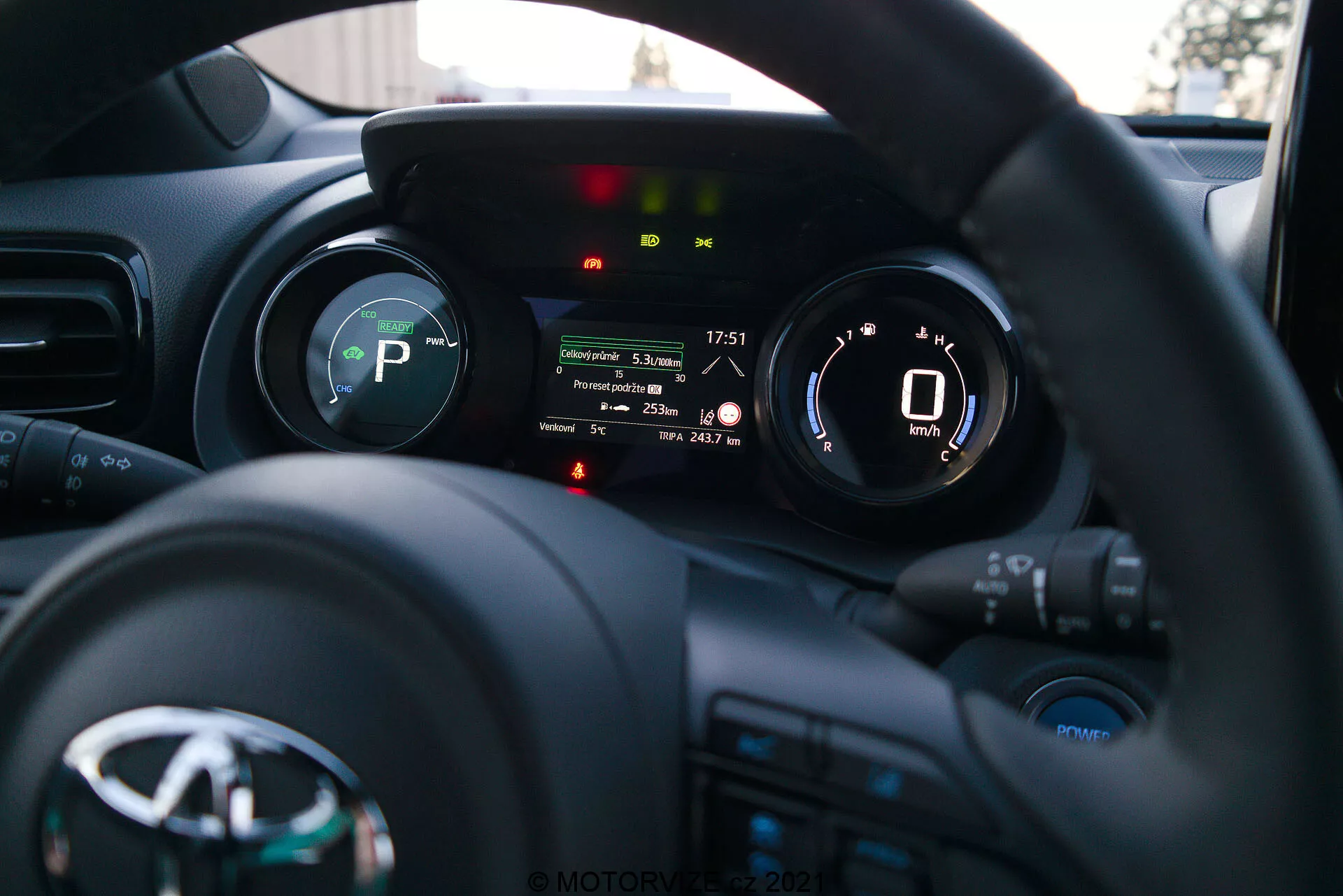 Une photographie de l'intérieur de la Toyota Yaris 2020 depuis la perspective du conducteur, mettant l'accent sur le volant et le tableau de bord. Le volant, avec le logo du fabricant et plusieurs boutons de commande pour les médias, la communication et le régulateur de vitesse, est en bas. Le tableau de bord derrière le volant présente un écran numérique affichant un compteur de vitesse indiquant 0 km/h et des indicateurs pour la marche (P pour Park), le rappel de ceinture de sécurité, l'alerte de porte ouverte et d'autres voyants d'état du véhicule. La faible profondeur de champ met en évidence le tableau de bord, tandis que le tableau de bord et l'intérieur de la voiture sont légèrement flous en arrière-plan. L'image, probablement prise en fin d'après-midi ou en début de soirée, montre une instrumentation moderne et une technologie d'interface du véhicule.