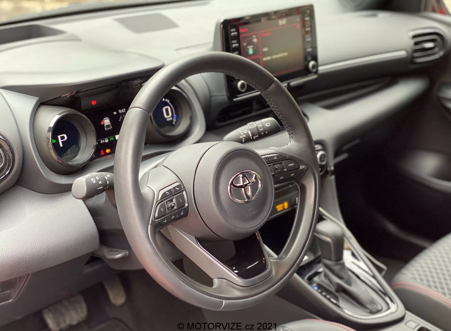 Vue intérieure d'une Toyota Yaris 2020 depuis le siège passager avant, se concentrant sur la zone du conducteur. L'image met en évidence un volant Toyota à trois branches avec des boutons de commande pour l'audio, le Bluetooth et le régulateur de vitesse, avec l'emblème Toyota au centre. Derrière le volant, le tableau de bord numérique est partiellement visible, indiquant que la voiture est en mode 'P' (parc). La console centrale montre l'écran d'infodivertissement et les commandes de la climatisation en dessous, ainsi que des ports USB ou auxiliaires possibles. L'image montre également un levier de vitesse automatique avec un design contemporain et une botte de type cuir. L'intérieur présente un design moderne avec une combinaison de matériaux doux au toucher et des accents argentés, ainsi que des porte-gobelets ou de petits compartiments de rangement à côté du levier de vitesse. Un panneau de porte partiellement visible révèle un panneau de commande des fenêtres, suggérant des fenêtres électriques.