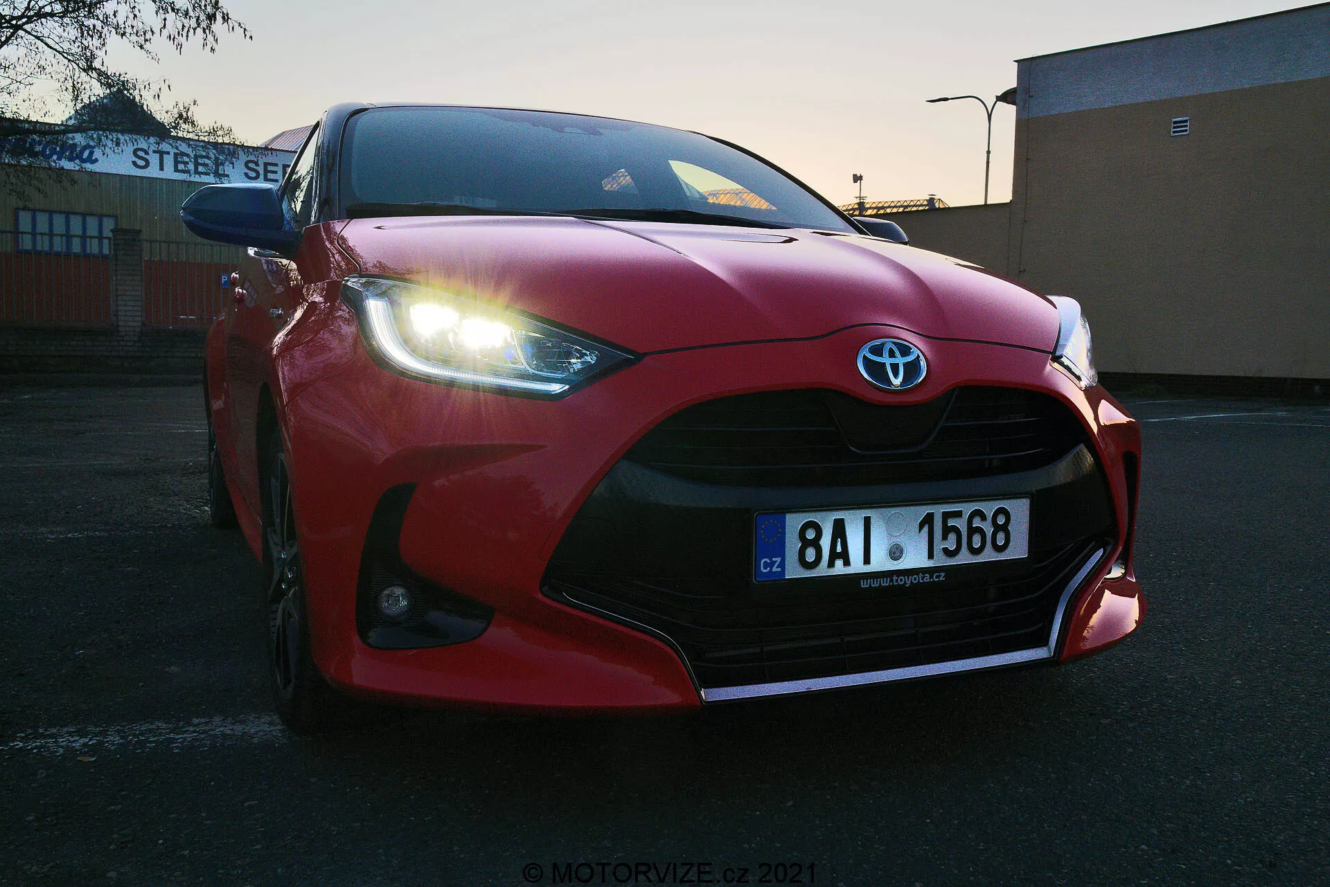 Gros plan en contre-plongée sur une Toyota Yaris 2020 rouge garée à l'extérieur au lever ou coucher du soleil. Grille avant, pare-chocs, capot sculpté aérodynamiquement, phares LED acérés et grandes prises d'air mettant en valeur le badge Toyota. Plaque d'immatriculation européenne visible, située sur un fond industriel ou commercial avec bâtiment 'STEEL SERVICE'. Ciel dégagé avec lumière dégradée mettant en avant le design sportif du véhicule.